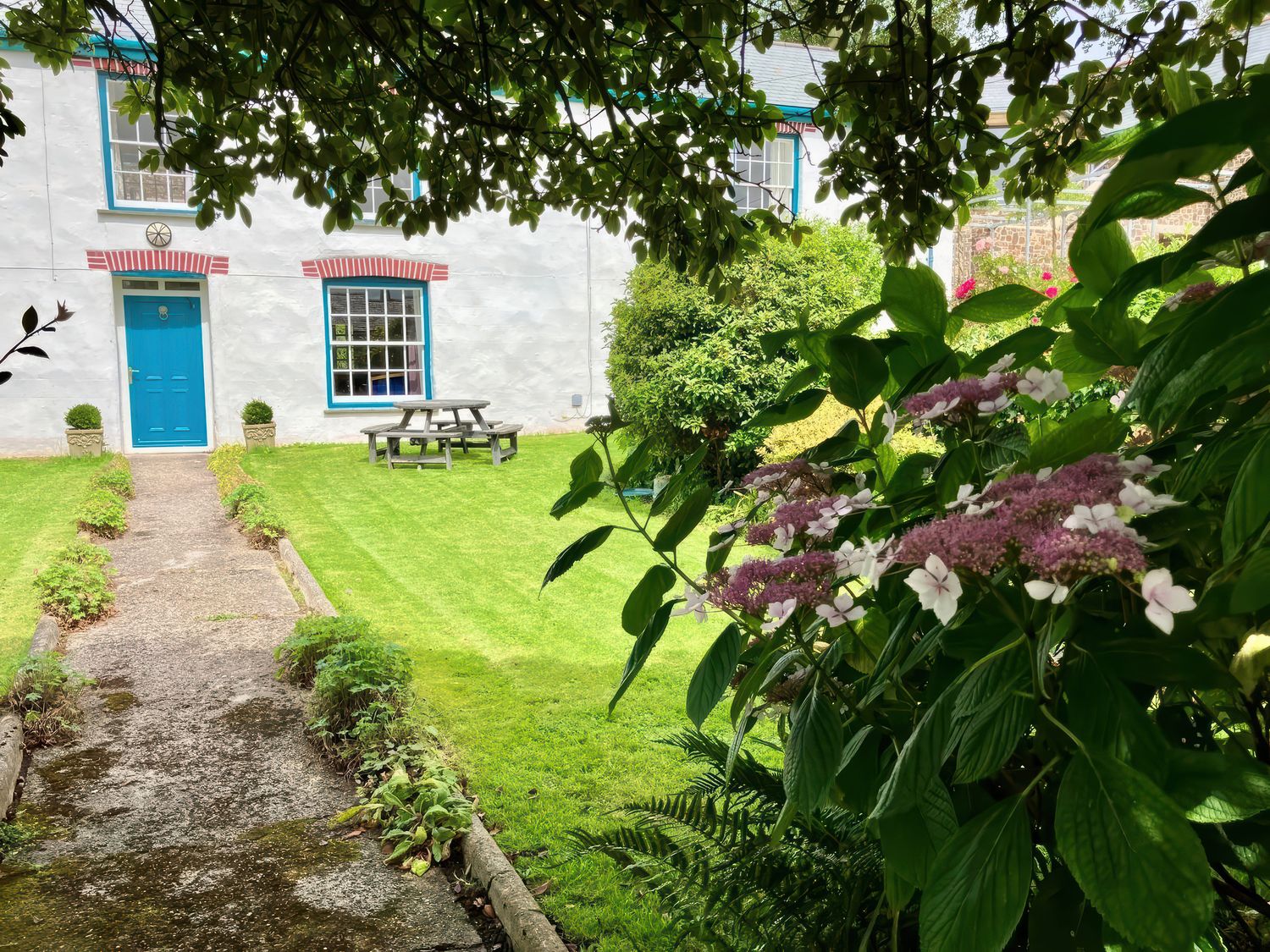 A garden with flowers and bushes leading to a house with a blue door and windows at Langford Barton in Marhamchurch near Widemouth Bay
