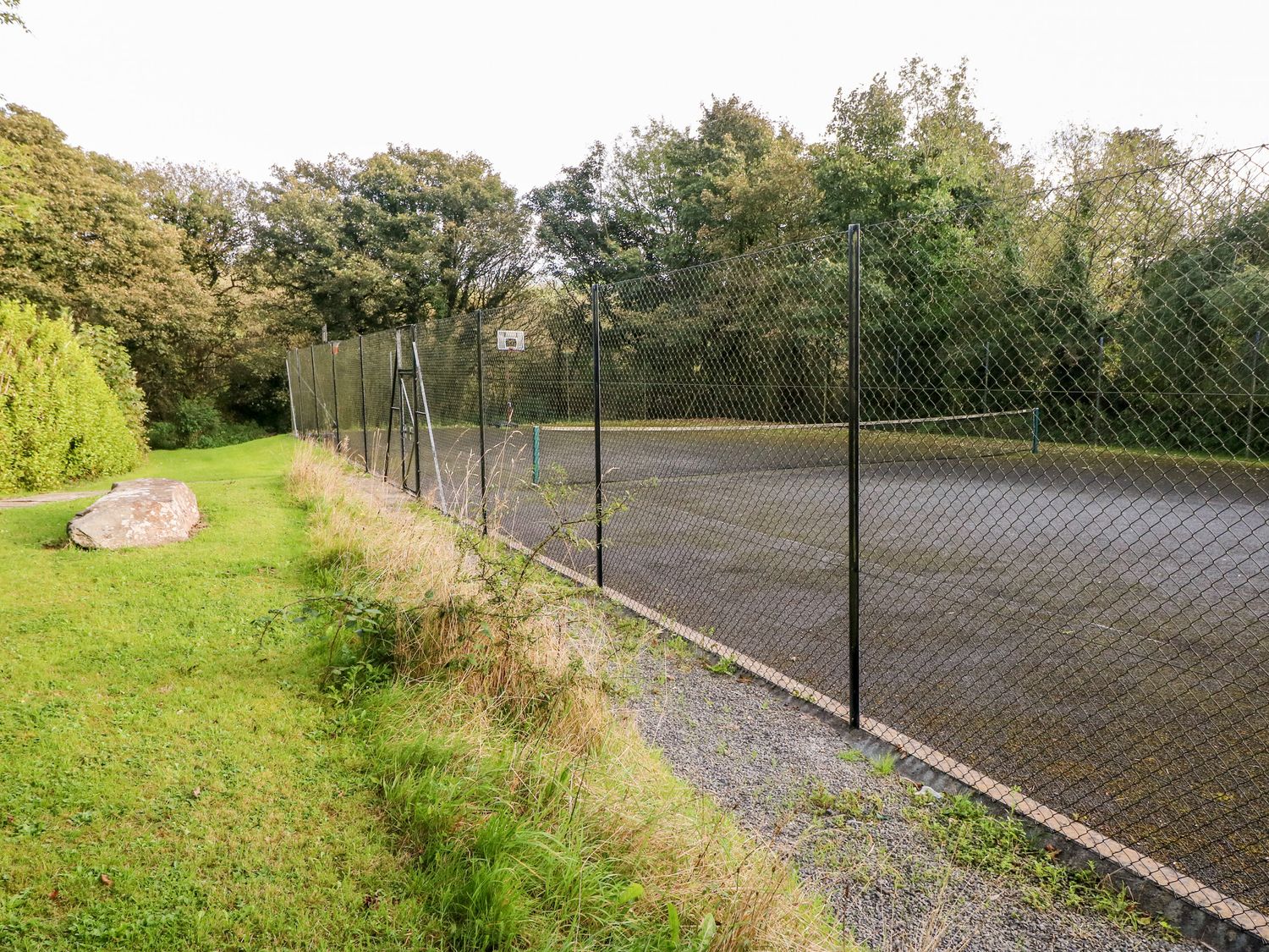 An outdoor tennis court with a chain link fence and a basketball hoop next to a grassy area with trees at Swallows Cottage in Maenclochog