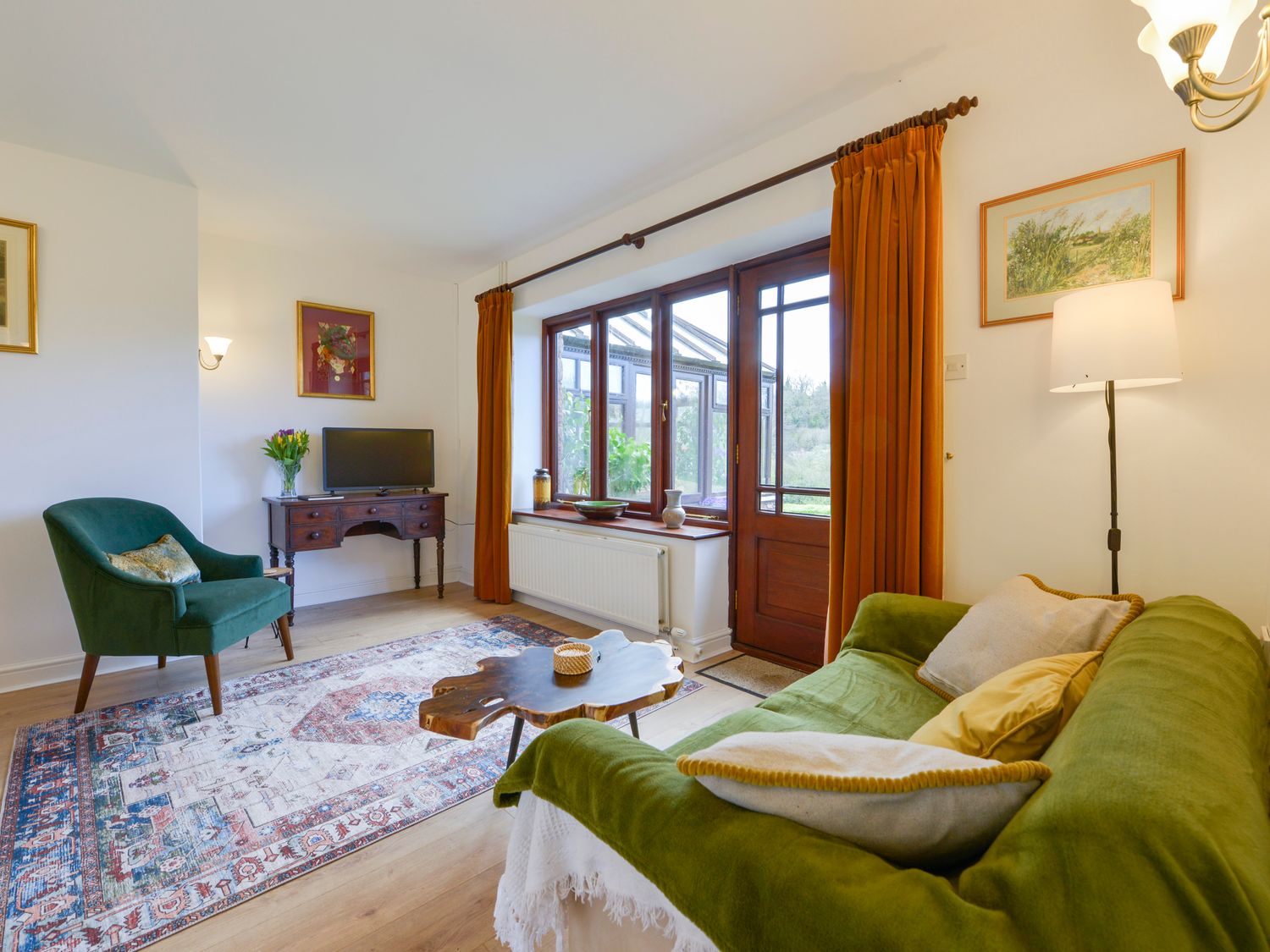 A living room with a green sofa, wooden coffee table, armchair, desk with TV, and large window with orange curtains at Little Barton in Stogumber near Watchet