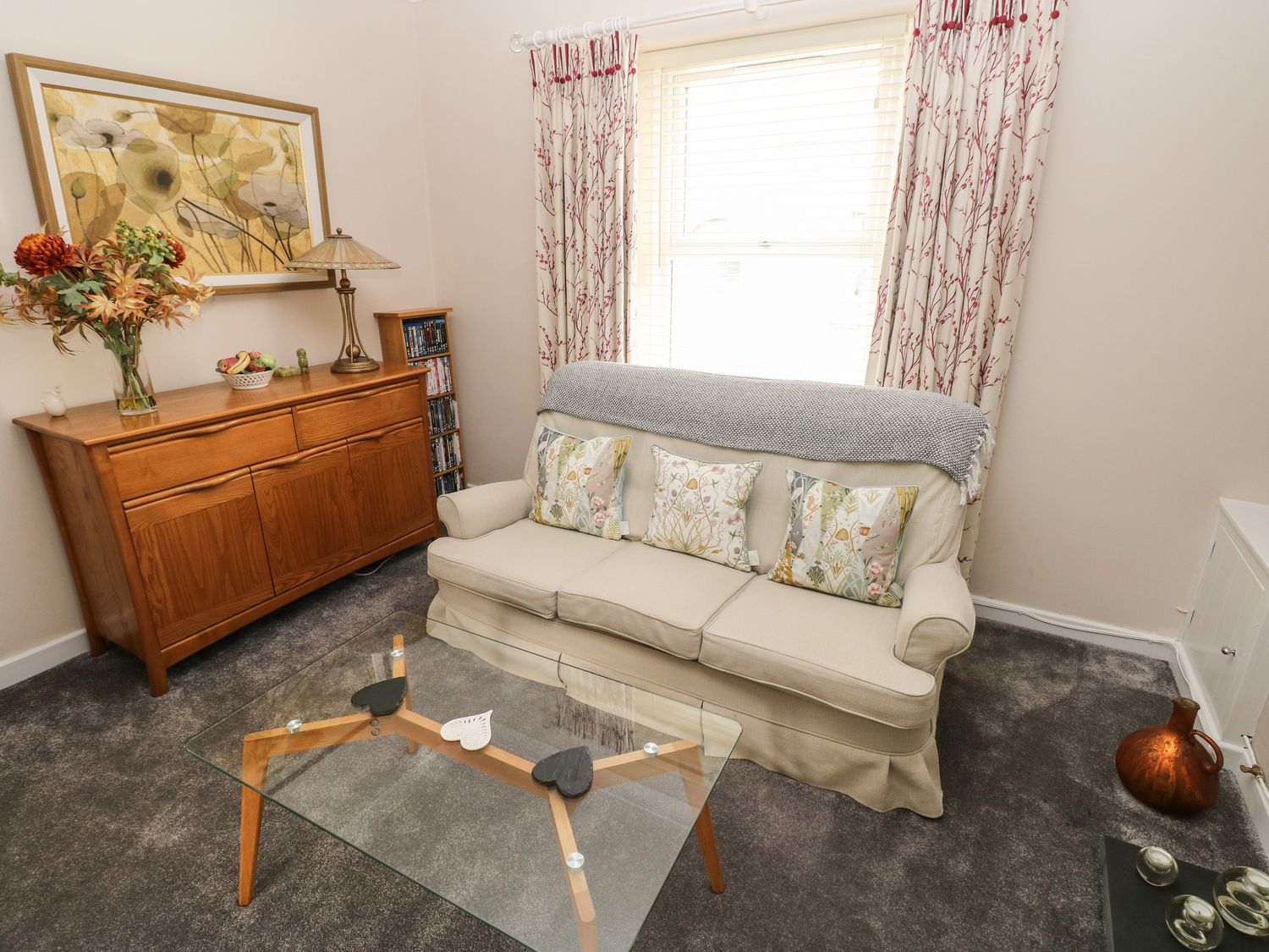A living room with a cream sofa and patterned cushions a glass coffee table wooden cabinet and decorative items at Holly Cottage in Pembroke