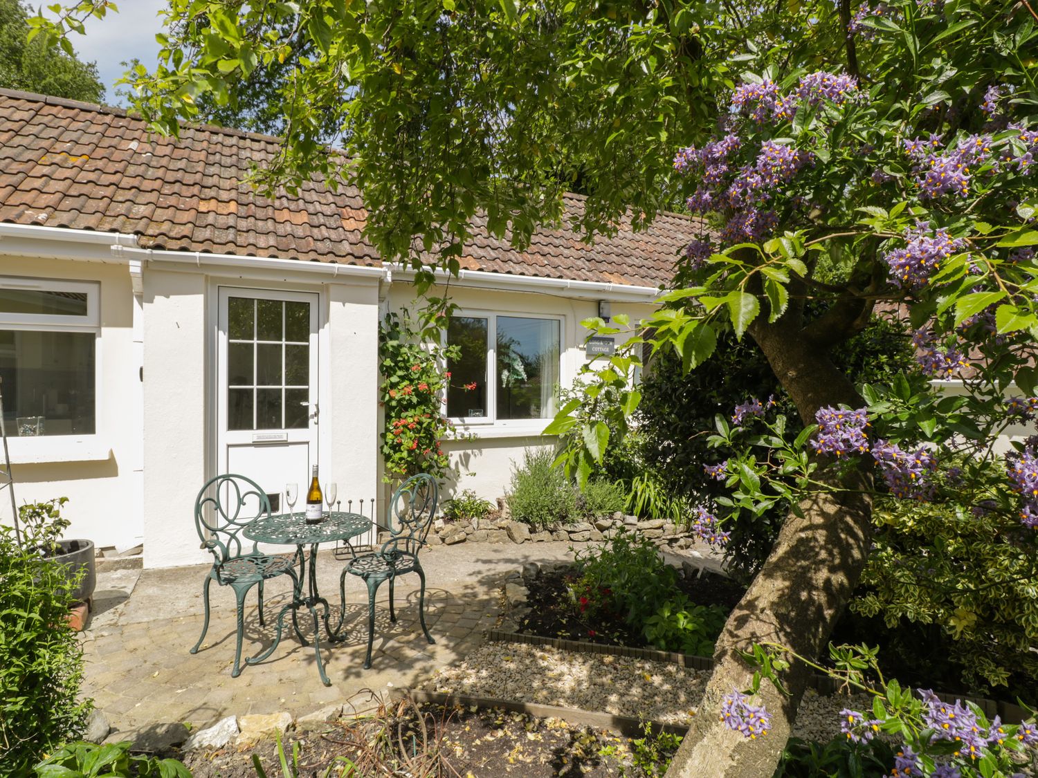 A patio with a metal table and two chairs outside a cottage with a garden and flowering tree at Long Batch Cottage in Ditcheat