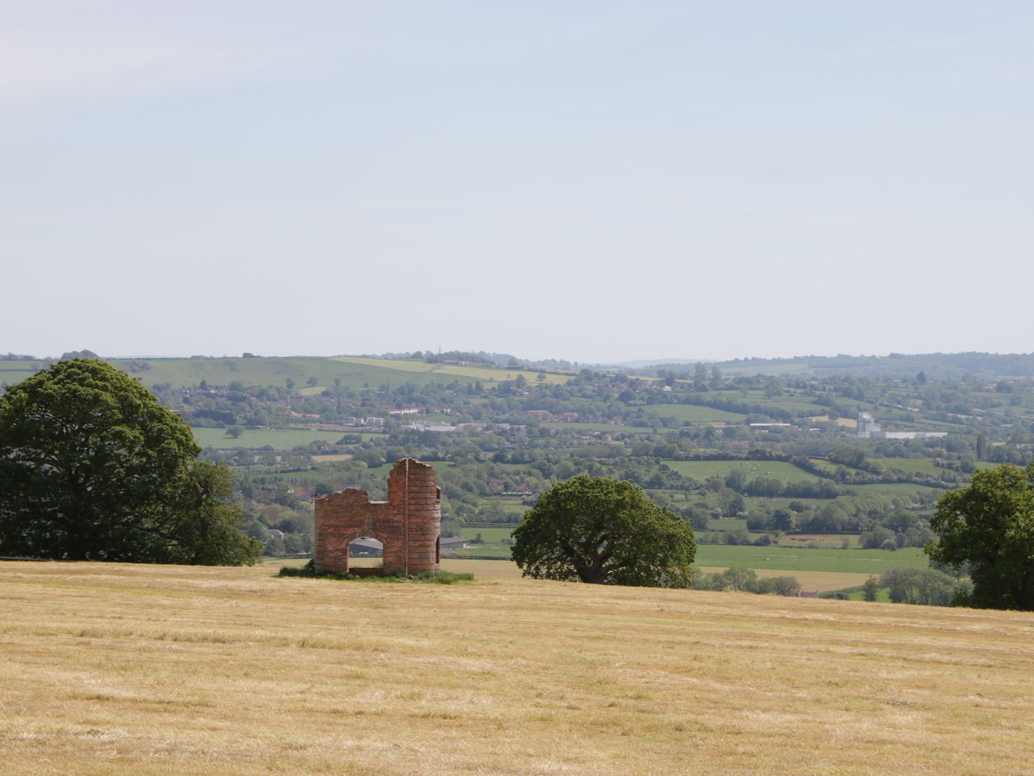 A grassy field with a small brick ruin surrounded by trees and hills in the distance at Long Batch Cottage in Ditcheat