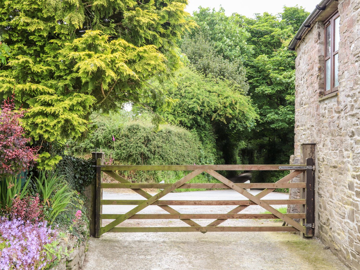 A wooden gate on a driveway next to a stone building with trees and bushes in the background at Keepers Cottage in Liskeard