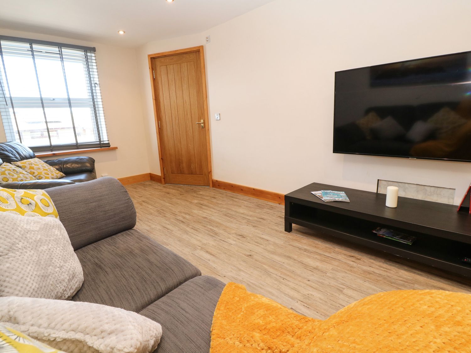 A living room with a gray sofa and cushions a black sofa a wooden door a large window with blinds a black TV stand with a candle and magazines and a wall-mounted TV at Godrevy Cottage in Hayle