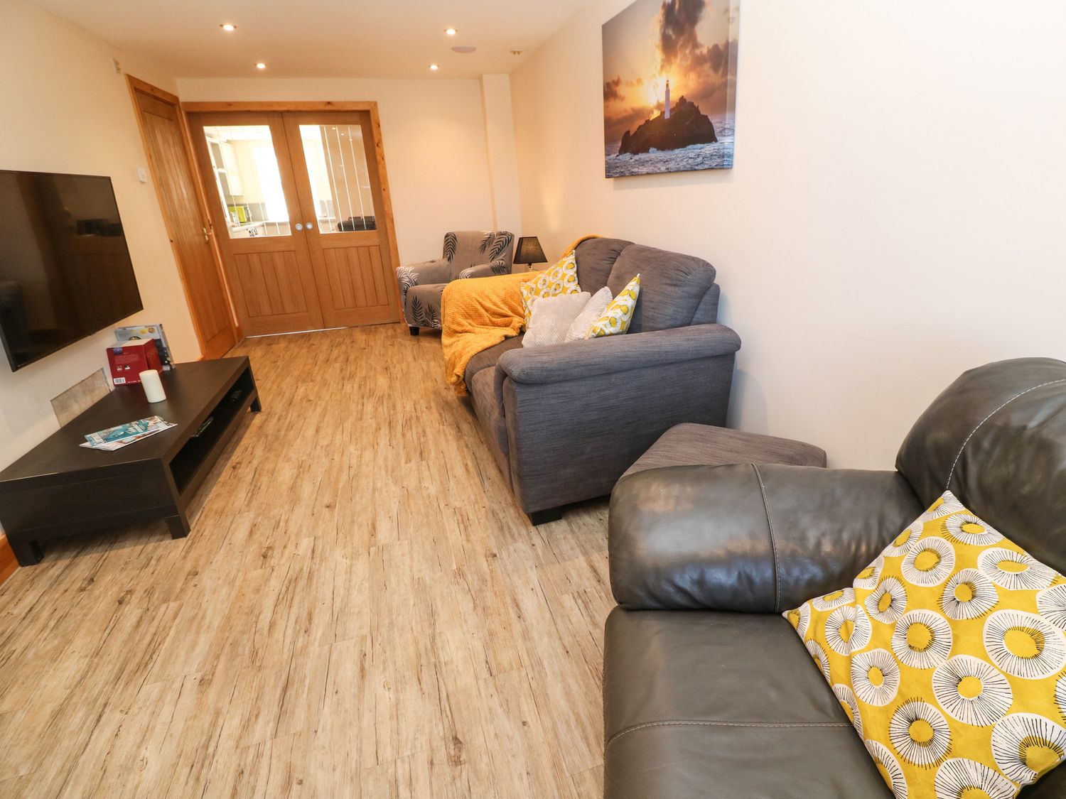 A living room with a gray sofa and armchair with yellow pillows a black coffee table and wooden double doors at Godrevy Cottage in Hayle