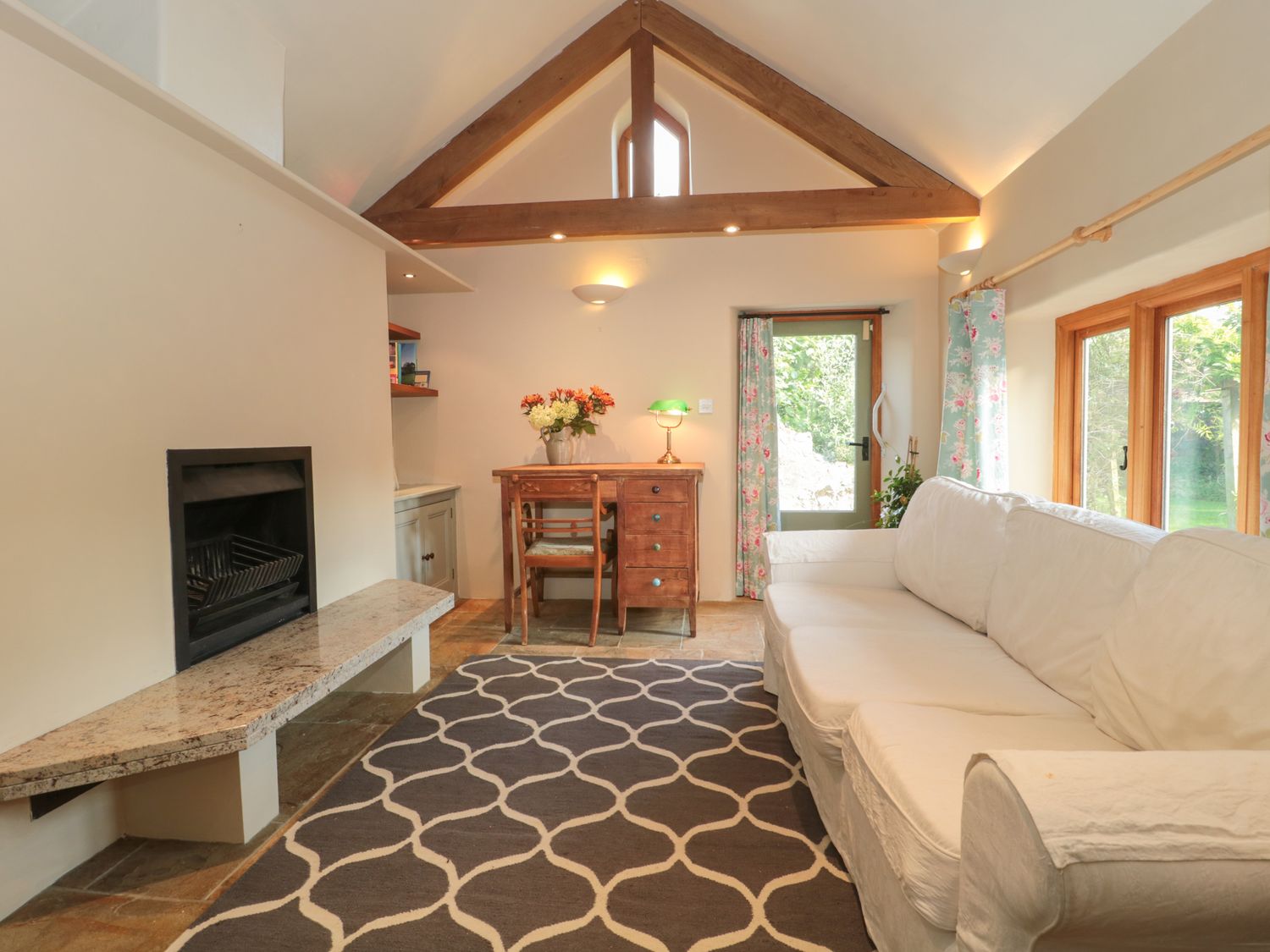 A living room with a white sofa a wooden desk with flowers and a lamp a fireplace and patterned rug at Wreath Green Annexe in Tatworth