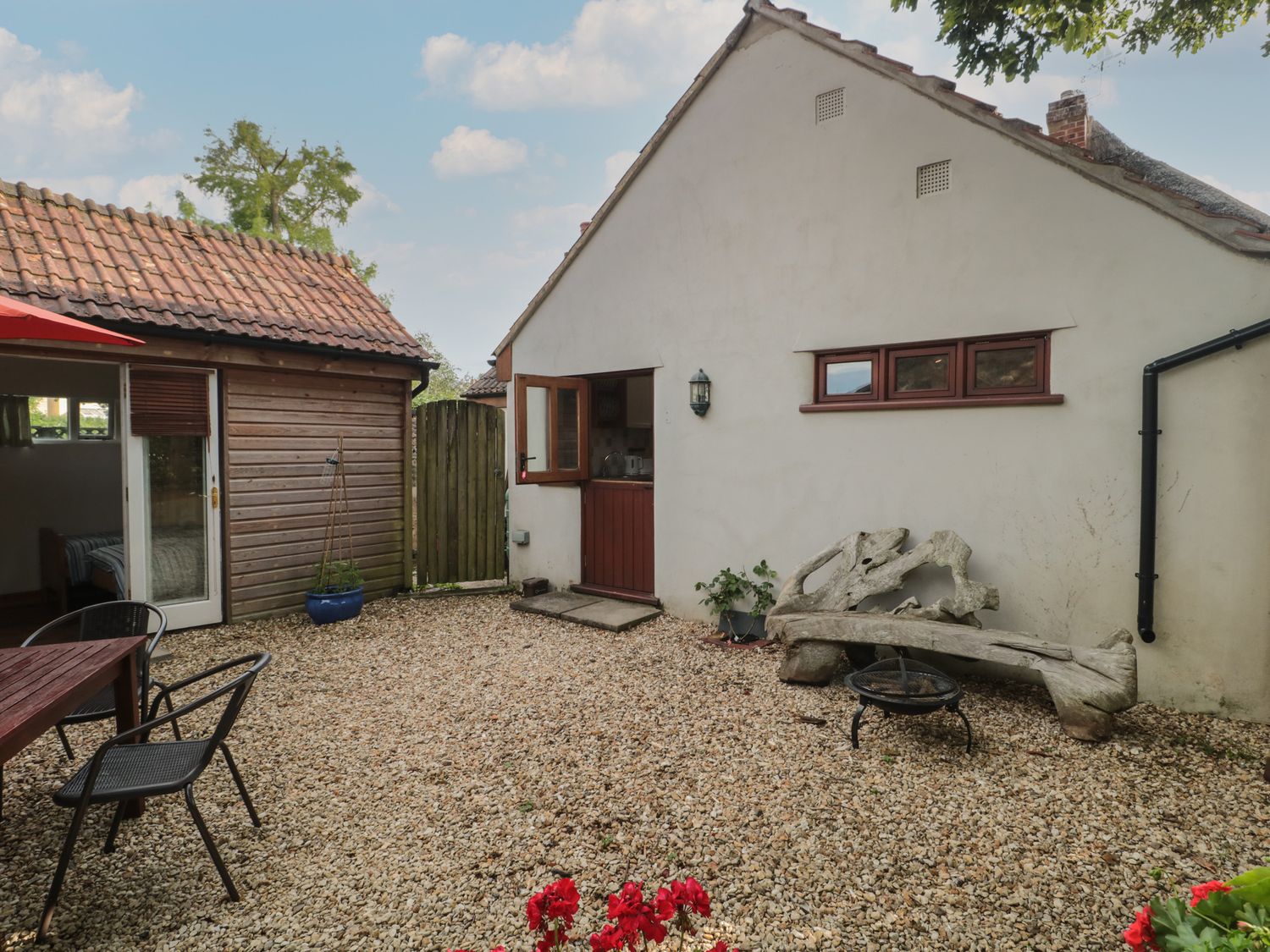 An outdoor patio area with gravel ground wooden bench metal fire pit table with chairs and two buildings at Wreath Green Annexe in Tatworth