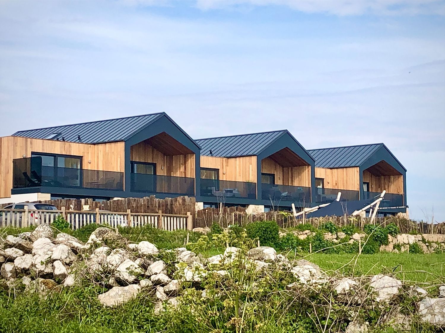 Three modern houses with wooden walls and dark roofs behind a stone and wooden fence in a green field at Sea Scape in Sennen