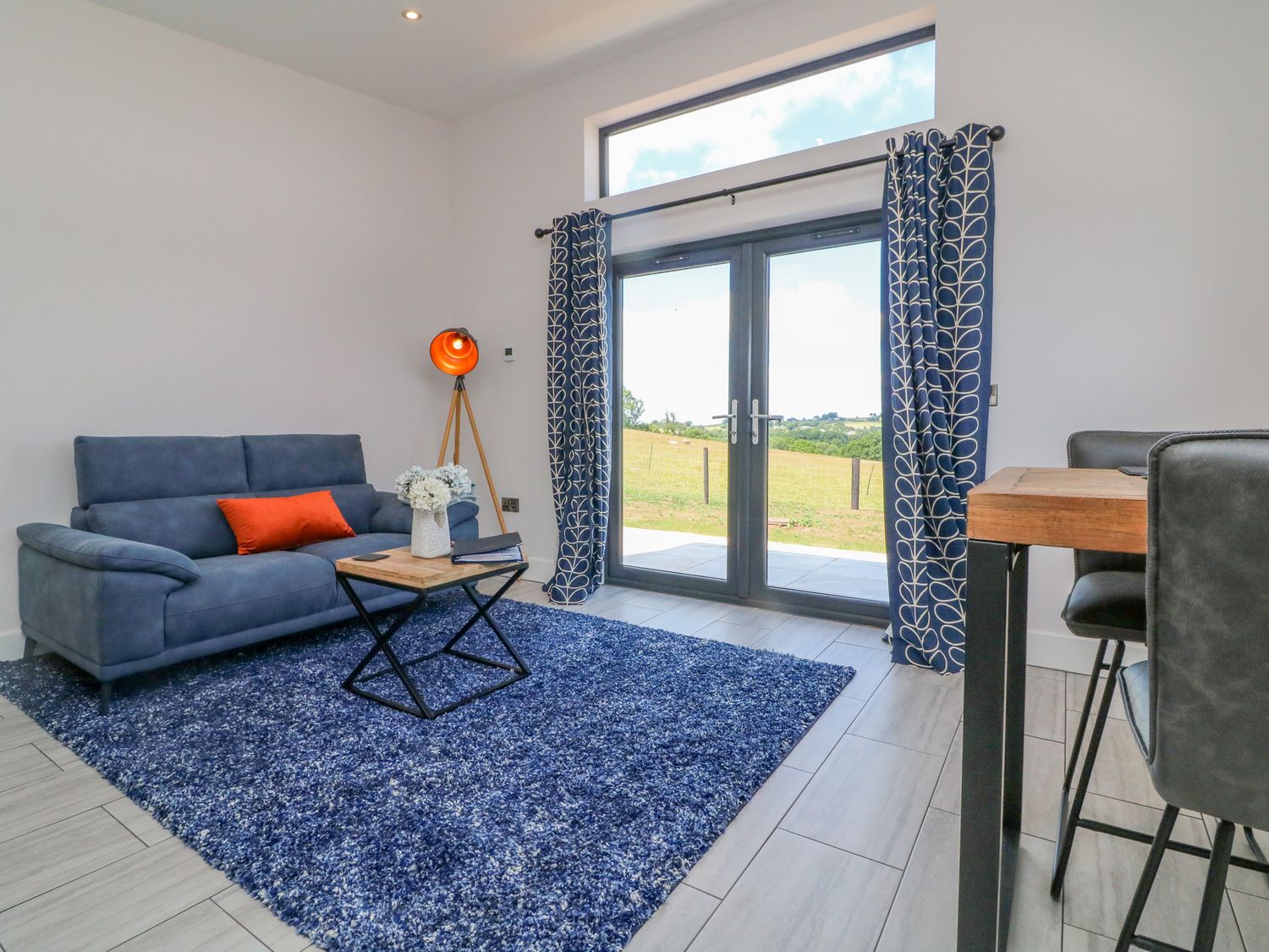 A living room with a blue sofa an orange cushion a floor lamp a wooden table blue patterned curtains and glass doors overlooking a field at The Cadeleigh in Cadeleigh near Tiverton