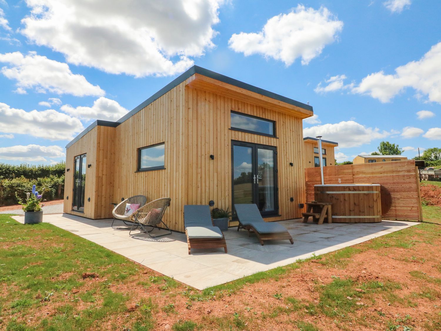 An outdoor patio area with lounge chairs a wooden hot tub and a modern wooden cabin at The Bickleigh in Cadeleigh near Tiverton