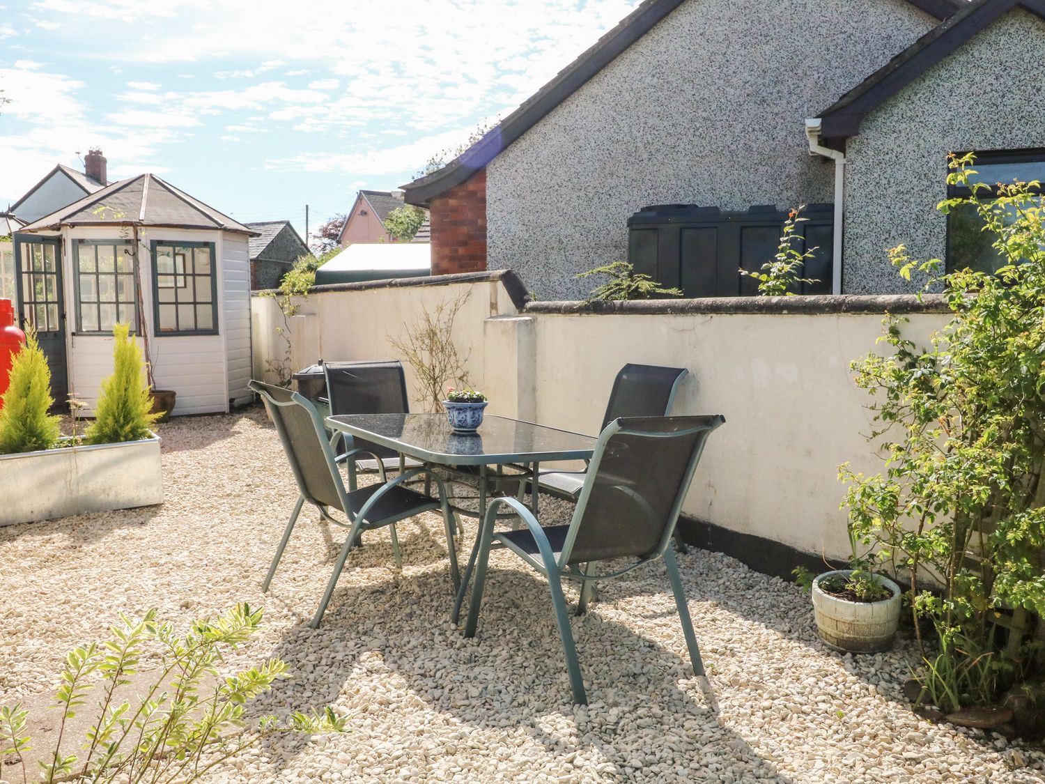 An outdoor patio area with a glass table and four chairs on gravel with a white shed and plants at Ash Tree Cottage in Bradworthy
