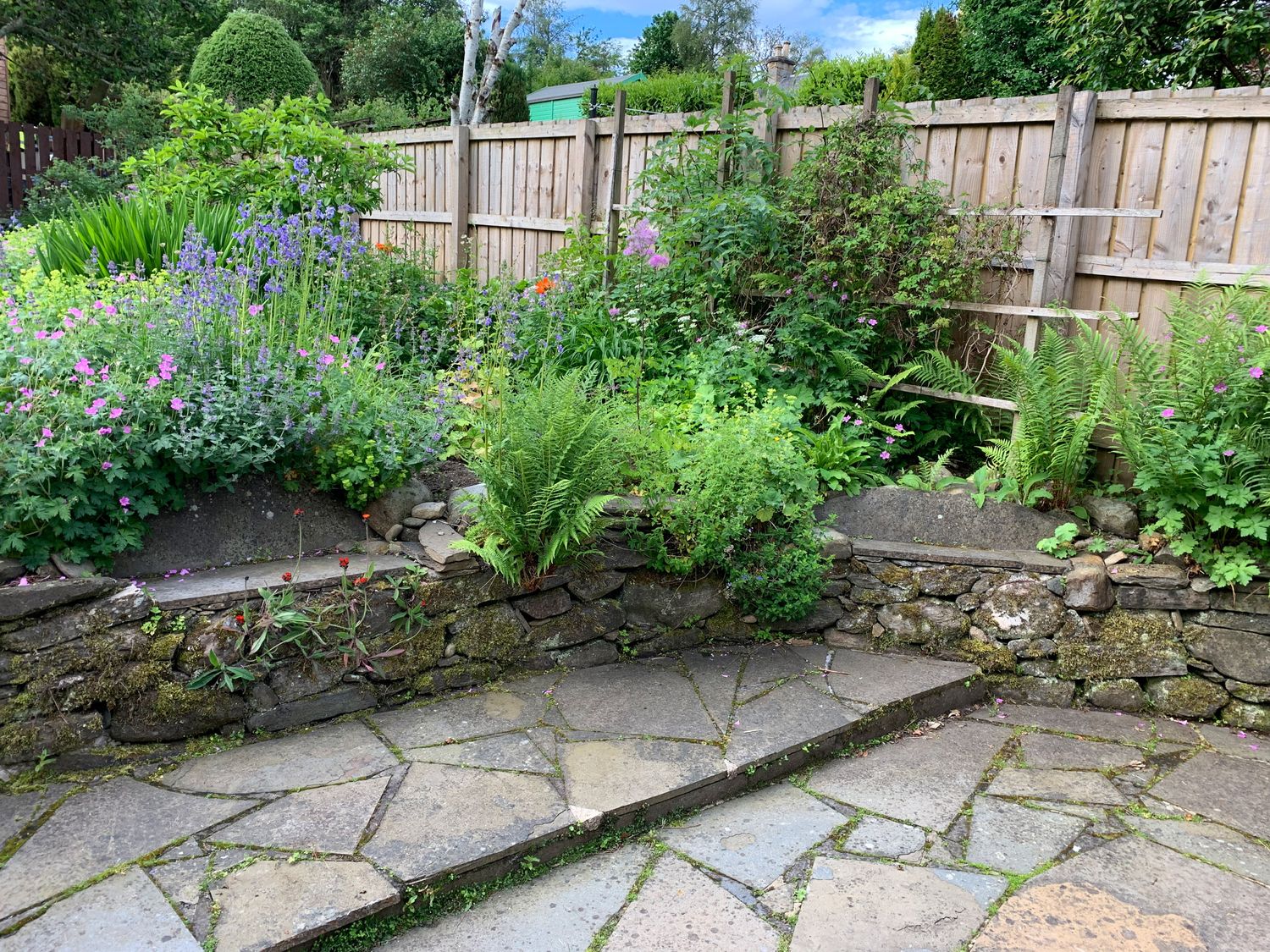 A garden area with stone paving and stone walls with plants and flowers behind a wooden fence at Old Schoolhouse in Grandtully
