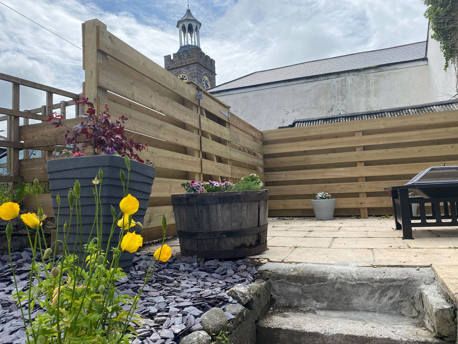 An outdoor patio area with flower pots wooden fencing and a clock tower in the background at Miner's Rest in St Day