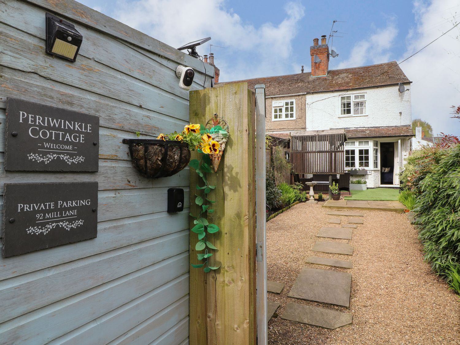 A garden path with stepping stones leading to a two-story cottage and a wooden fence with signs at Periwinkle Cottage in Barrow Upon Soar