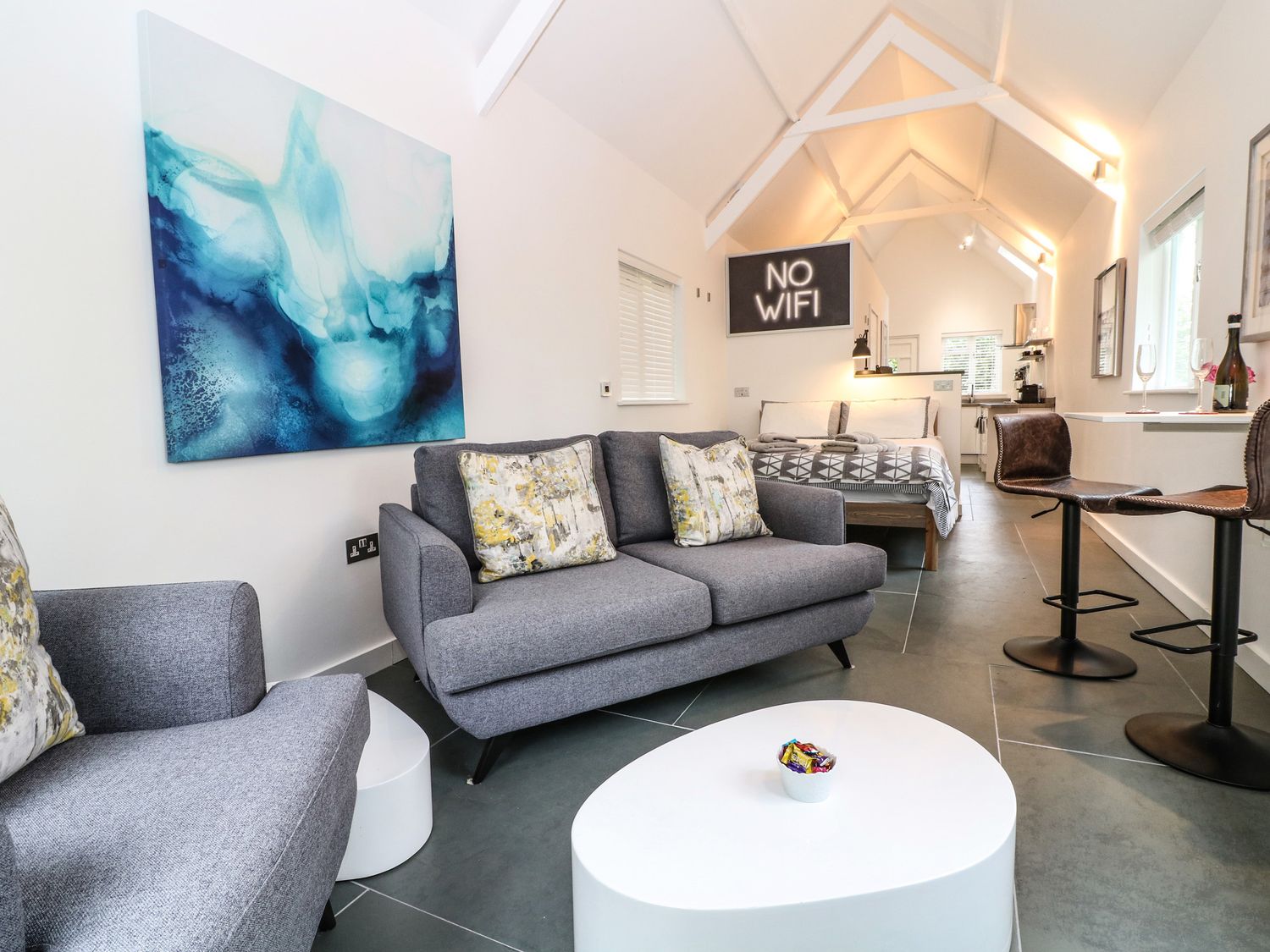 A living area with gray sofas, a white coffee table, bar stools, a bed, and a no wifi sign at Twigs in Woody Bay