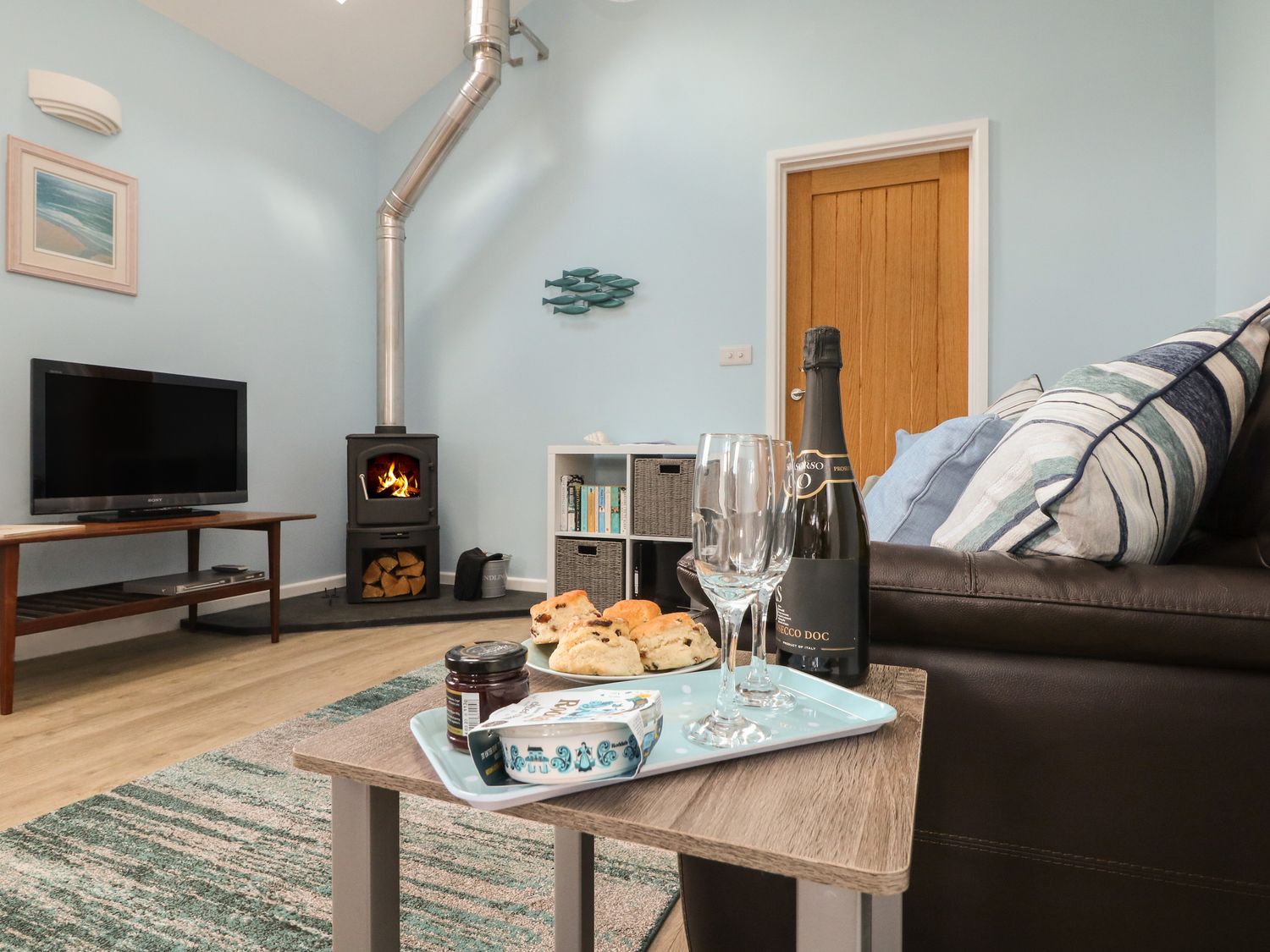 A living room with a wood stove television and a table with scones jam and a bottle of prosecco at Driftwood in Kelynack near St Just