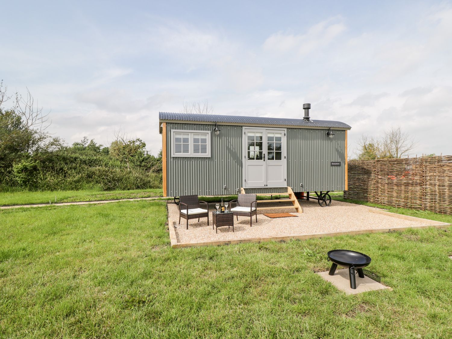 A small grey corrugated metal cabin with double doors and windows on a gravel patio with two chairs and a table on grass at Ridgeway in Uffington