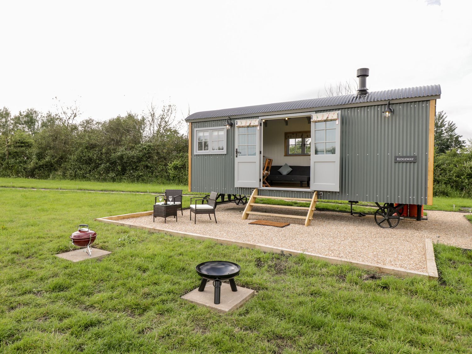 A small corrugated metal cabin on wheels with open double doors two chairs and a table outside a red kettle barbecue and a fire pit in a grassy area at Ridgeway Uffington
