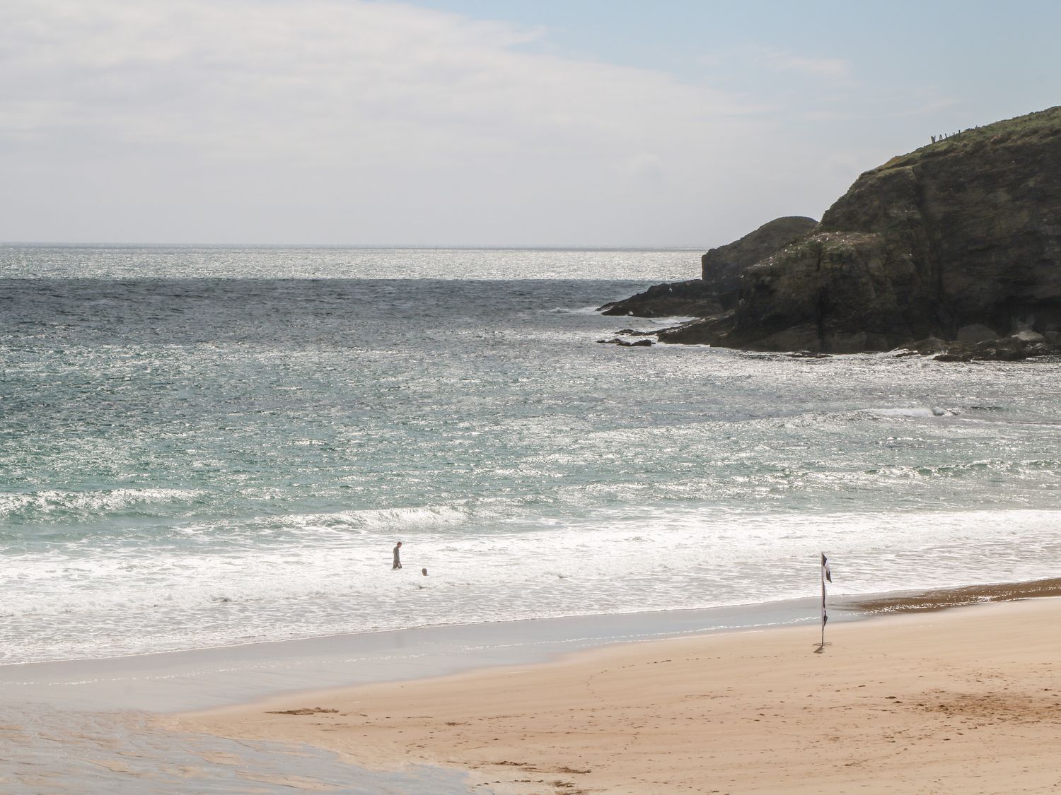 A beach with two people in the water a flag planted in the sand and rocky cliffs at Sandbanks in Praa Sands