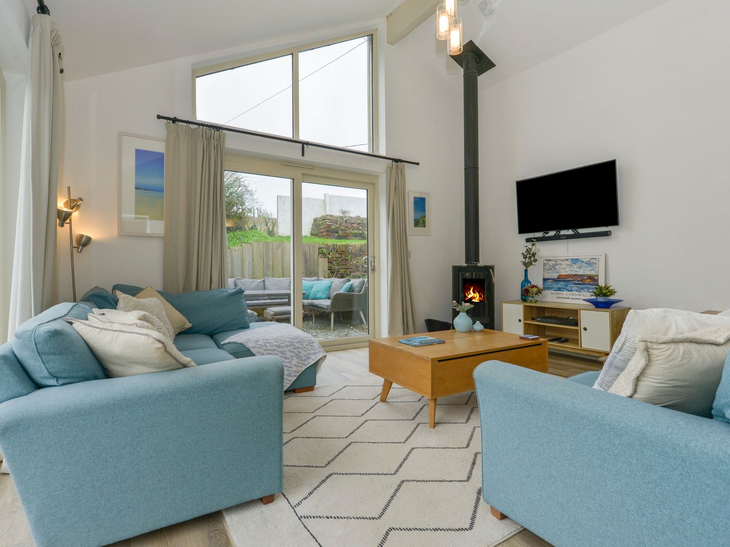 A living room with two blue sofas a wooden coffee table a wood-burning stove and a wall-mounted TV at Kelmscot Trelights