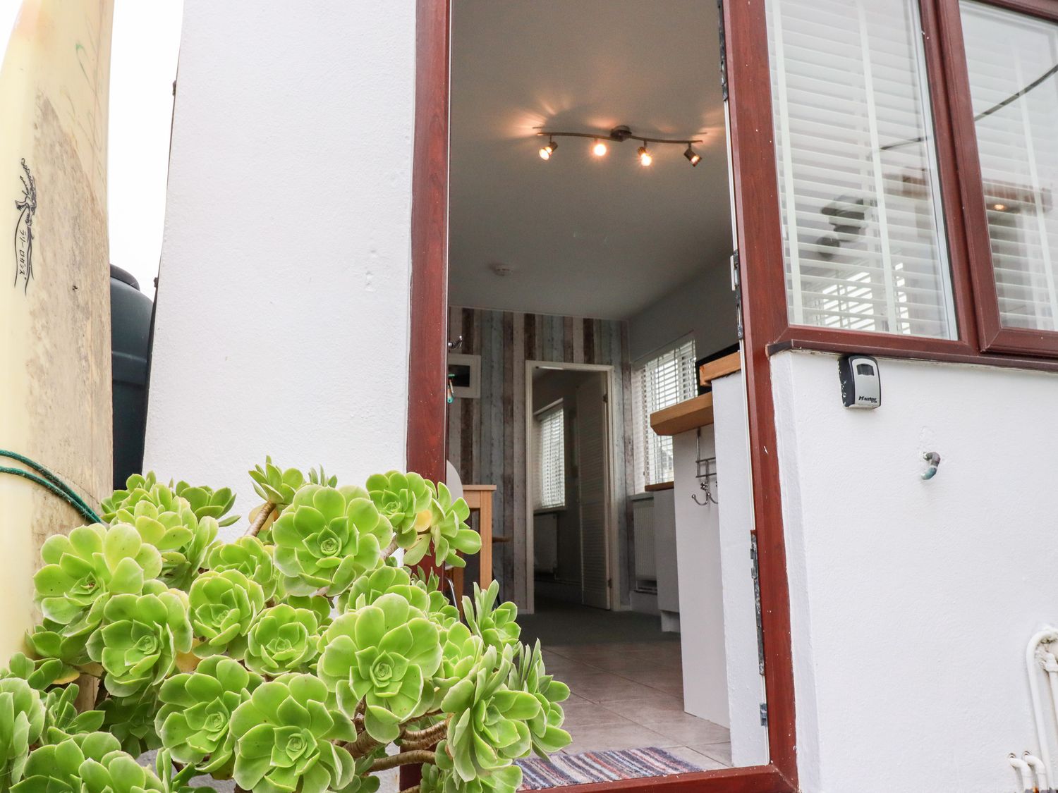 An open door leading into a room with tiled floor and wooden accent wall with a plant and surfboard outside at Endless Summer in Newquay