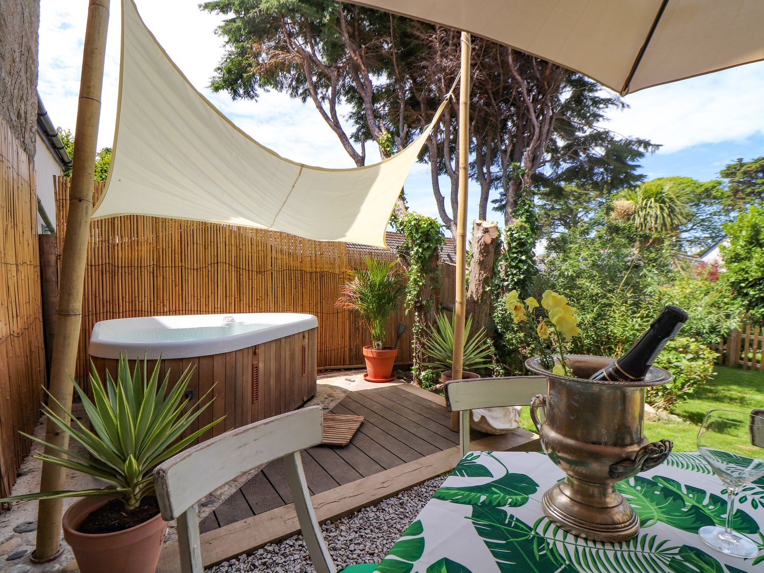An outdoor patio area with a hot tub shaded by a white sail and a table with a bottle in an ice bucket at Roncon's Villa in Goldsithney