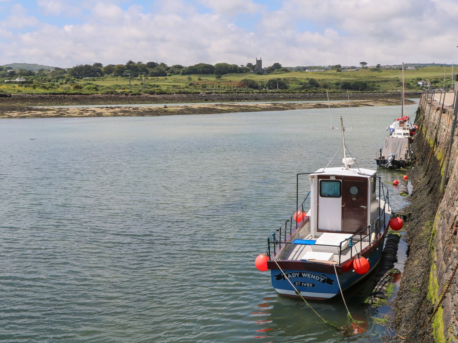A boat named Lady Wendy moored along a stone wall in a body of water with green fields and a church in the background at Primrose Cottage in Hayle