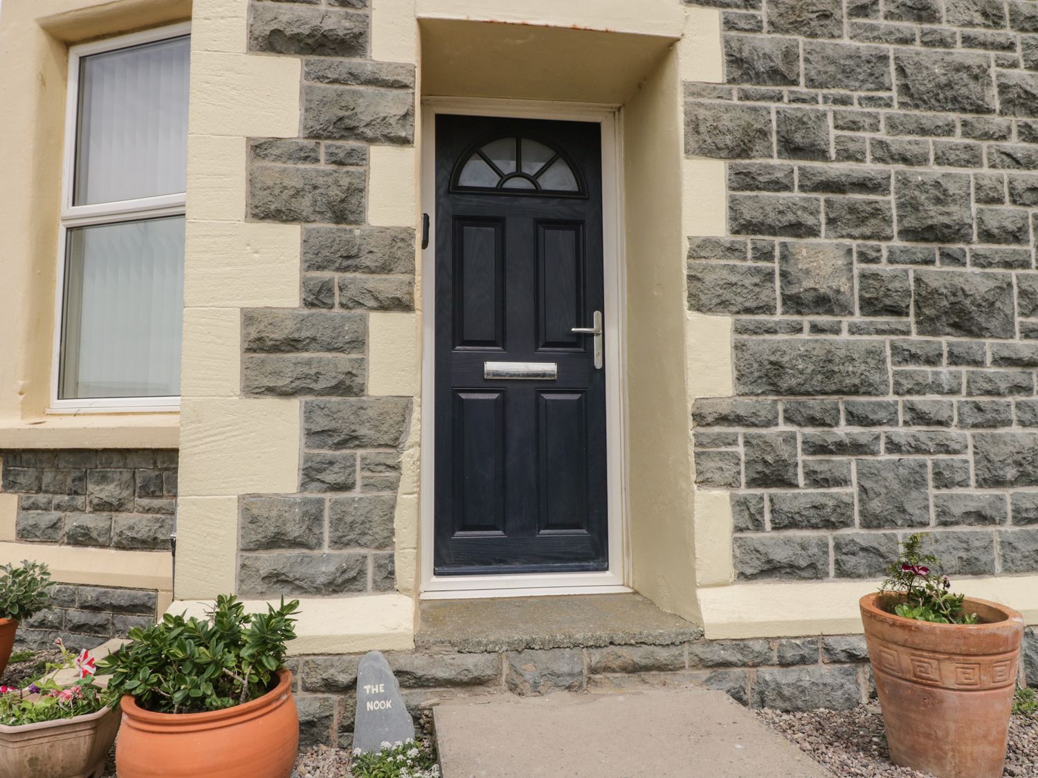 A black front door with a letterbox in a stone wall with flower pots on either side at The Nook in Tywyn