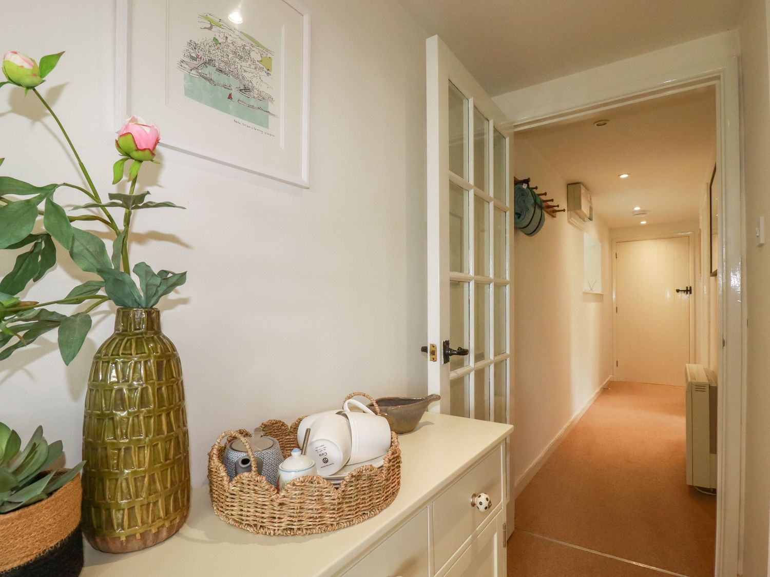 A hallway with a side table holding a green vase with flowers and a wicker basket with tea items at Bray Cottage in St Wenn near Winnard's Perch