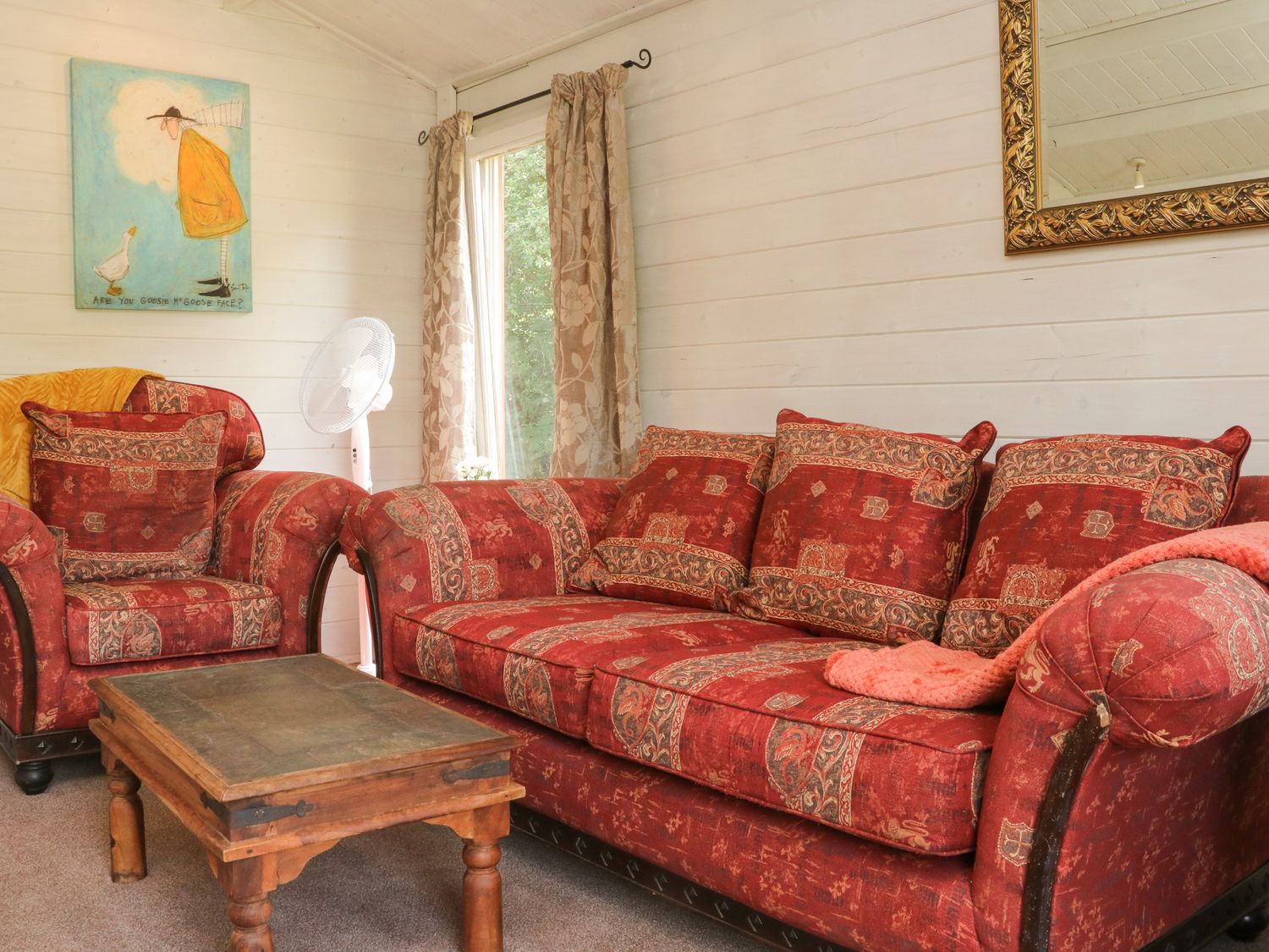 A living room with a patterned red sofa and armchair a wooden coffee table a standing fan curtains and a painting at The Cabin, Lowley Brook Farm Rezare near Launceston