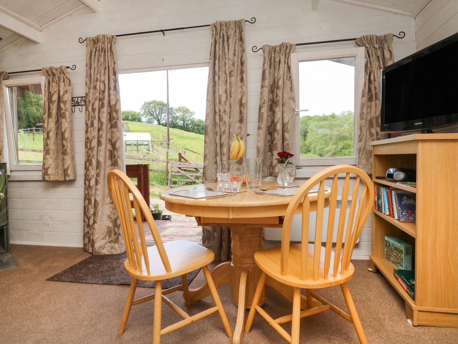 A dining area with a round wooden table and two chairs near windows with curtains and a wooden bookshelf with a TV at The Cabin Lowley Brook Farm Rezare near Launceston