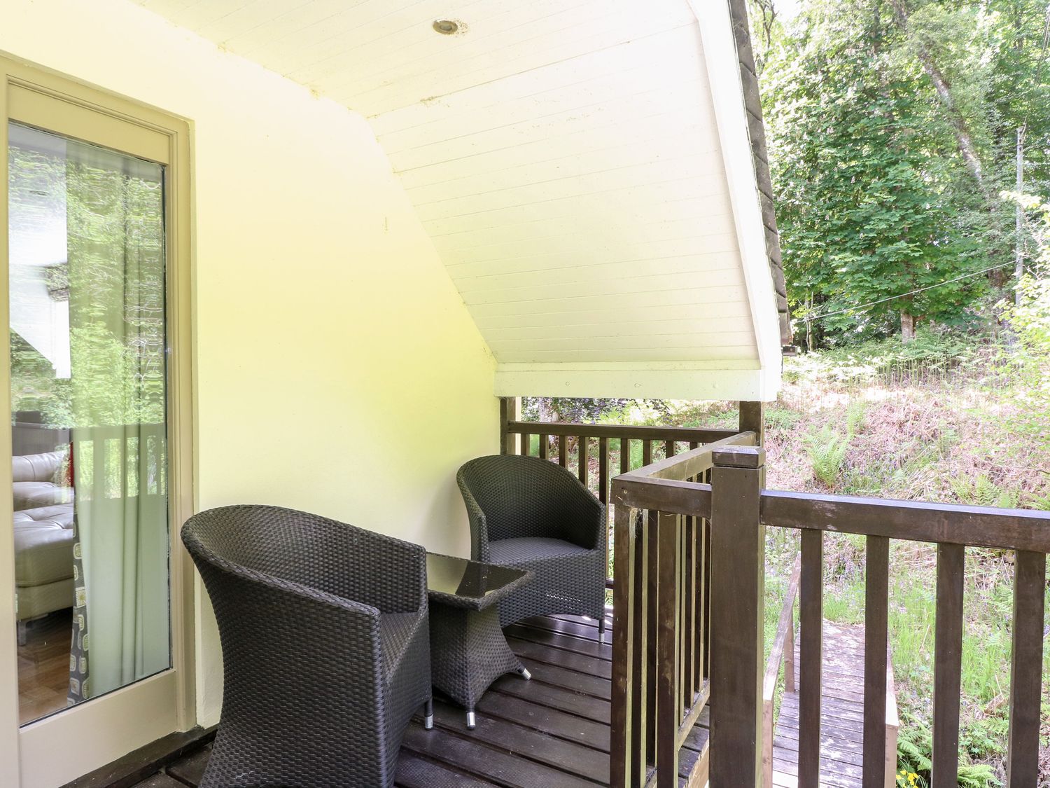 A small balcony with two wicker chairs and a glass top table overlooking greenery at Waterfall Apartment in Bearnock near Cannich