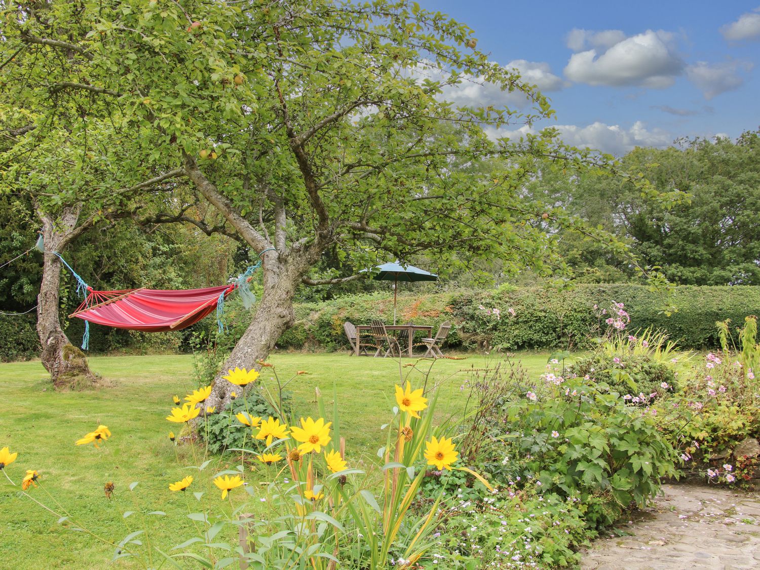 A garden with a red hammock between trees yellow flowers and a table with chairs under an umbrella at Denbigh Hall in Presteigne