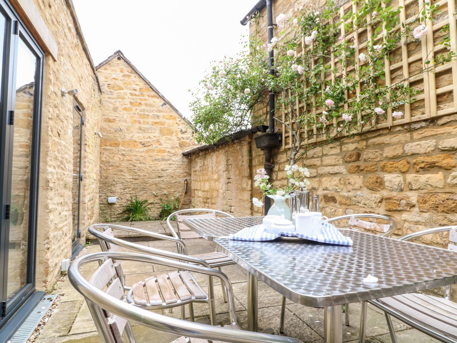An outdoor patio with metal table and chairs stone walls and climbing plants on a trellis at Ruskin House in Moreton-In-Marsh