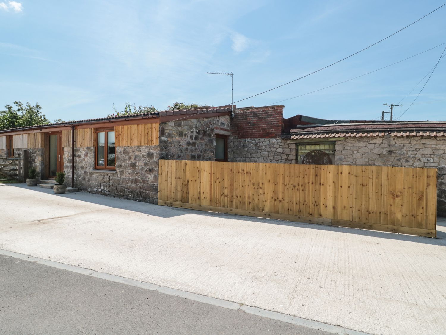 An exterior view of a stone and wood building with a wooden fence and concrete driveway at The Old Cow Shed in Banwell