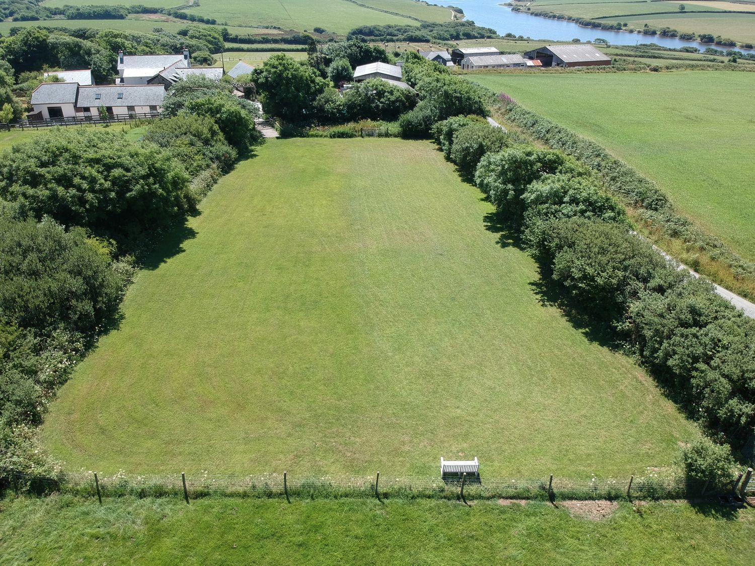 A large rectangular grass field bordered by trees with houses in the background at Woodpecker Cottage in Bradworthy near Bude