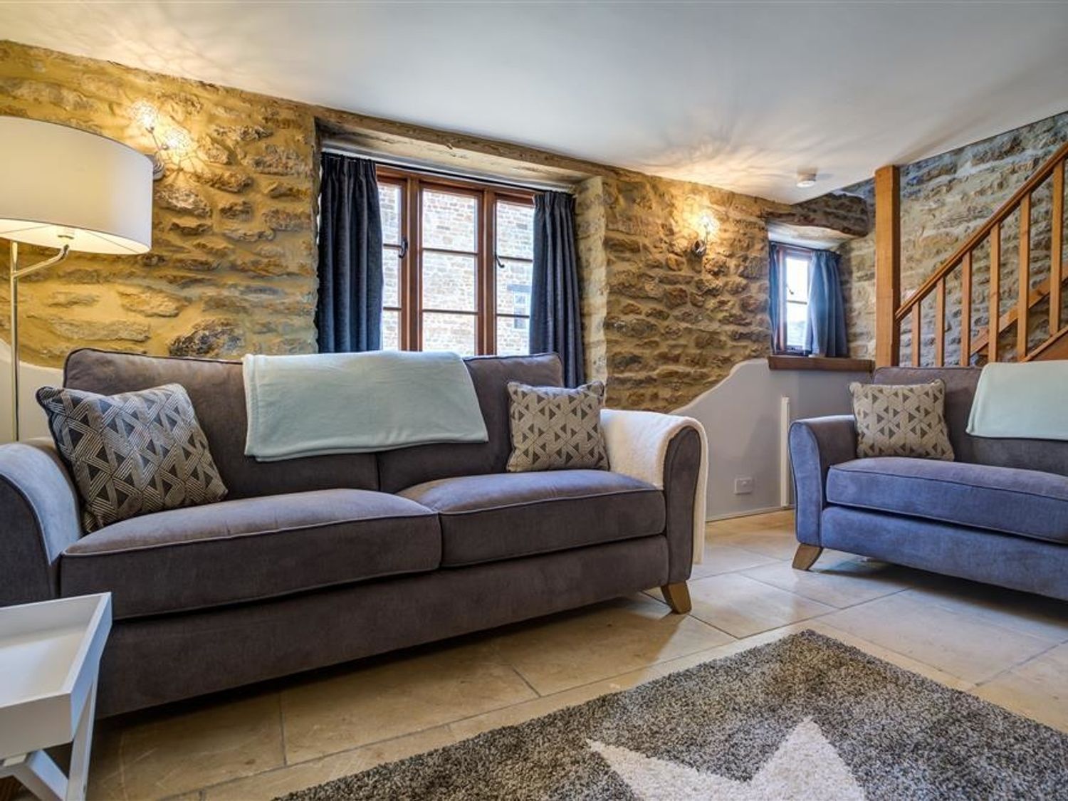 A living room with two gray sofas stone walls wooden window frames and a staircase at Hollytree Cottage in Hook Norton