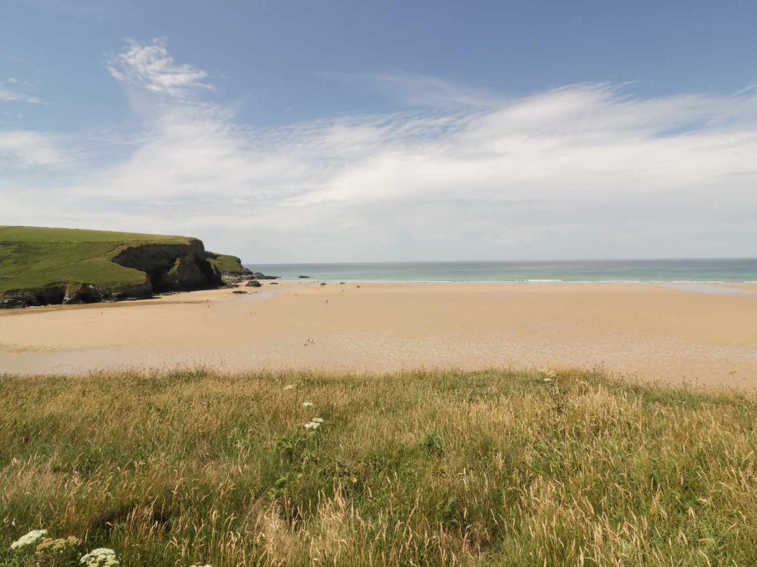 A sandy beach with a grassy hill in the foreground and a rocky cliff to the left at Woodlands Close in Padstow