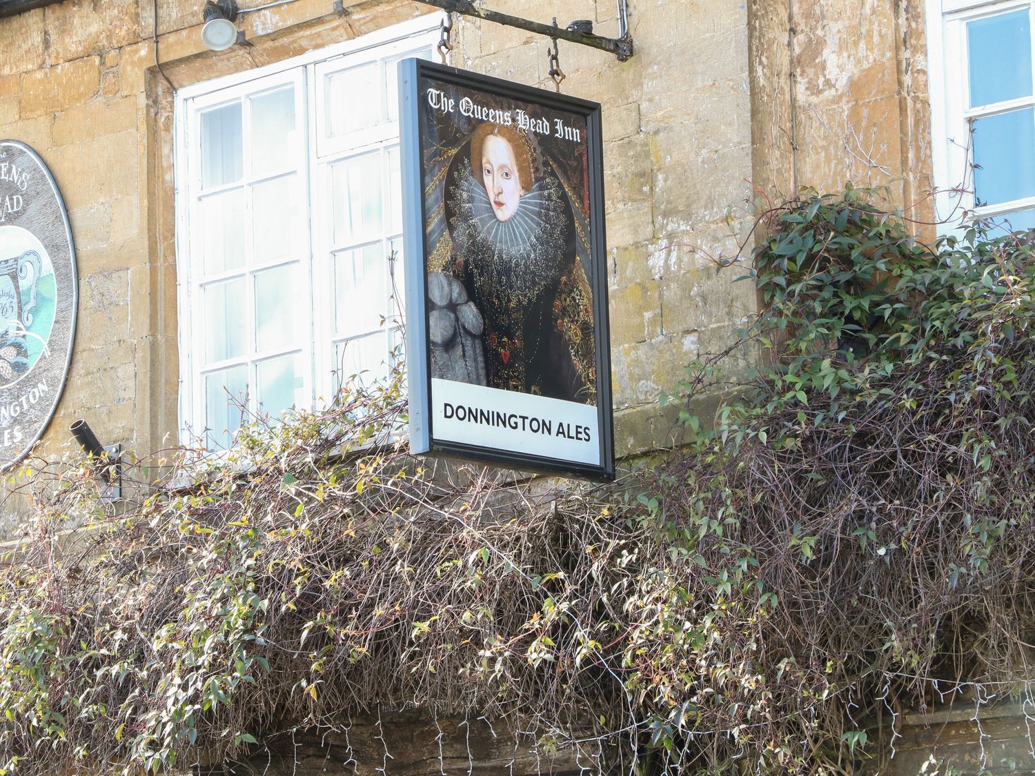 A pub sign with a portrait of a queen and text the queens head inn donnington ales on a stone building with windows and climbing plants