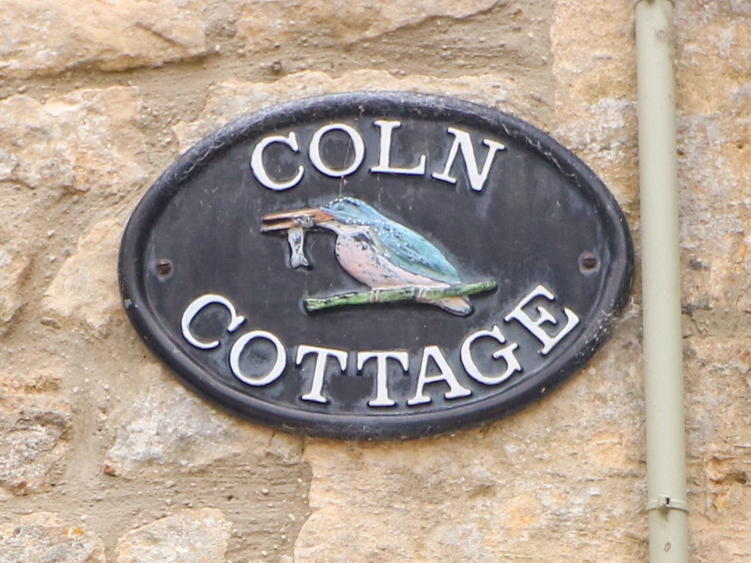 A wall-mounted oval plaque with a bird holding a fish and text reading Coln Cottage on a stone wall at Coln Cottage in Stow-On-The-Wold