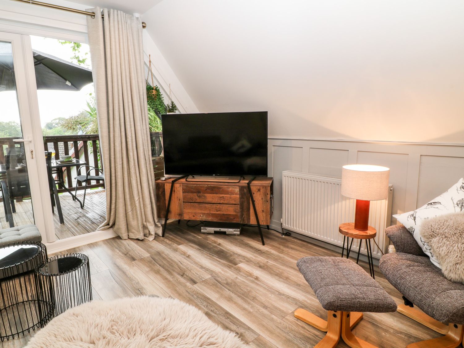 A living room with a television on a wooden stand a lamp on a side table and a chair with ottoman at Lavender Lodge in St Ann's Chapel Cornwall
