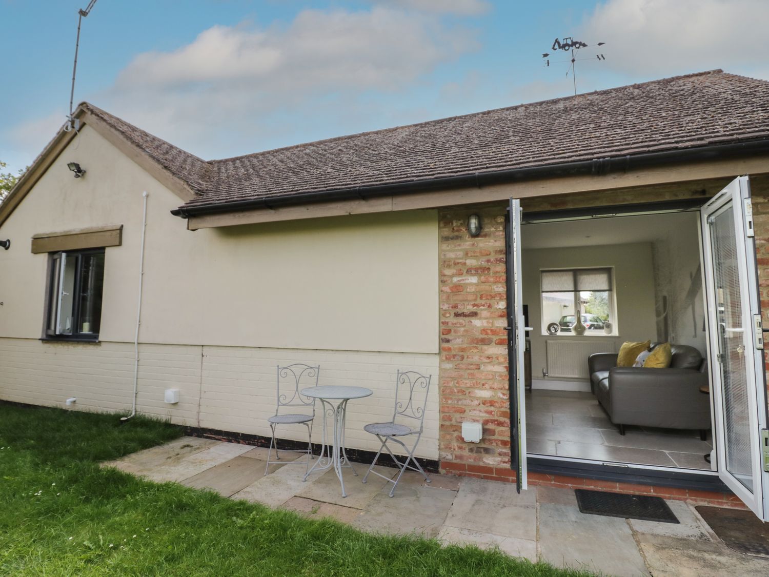 A patio with two metal chairs and a round table outside a single story house with open doors showing a living room with a sofa at Garden Cottage in Upton Upon Severn