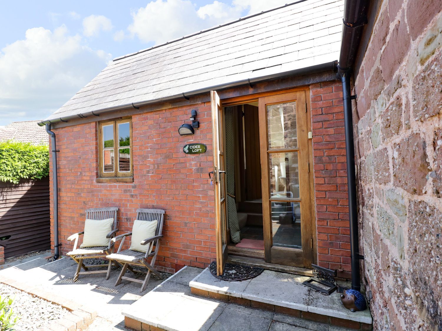 A small brick building with a tiled roof wooden chairs with cushions outside and a wooden door open at The Cider Loft in Whitchurch Herefordshire