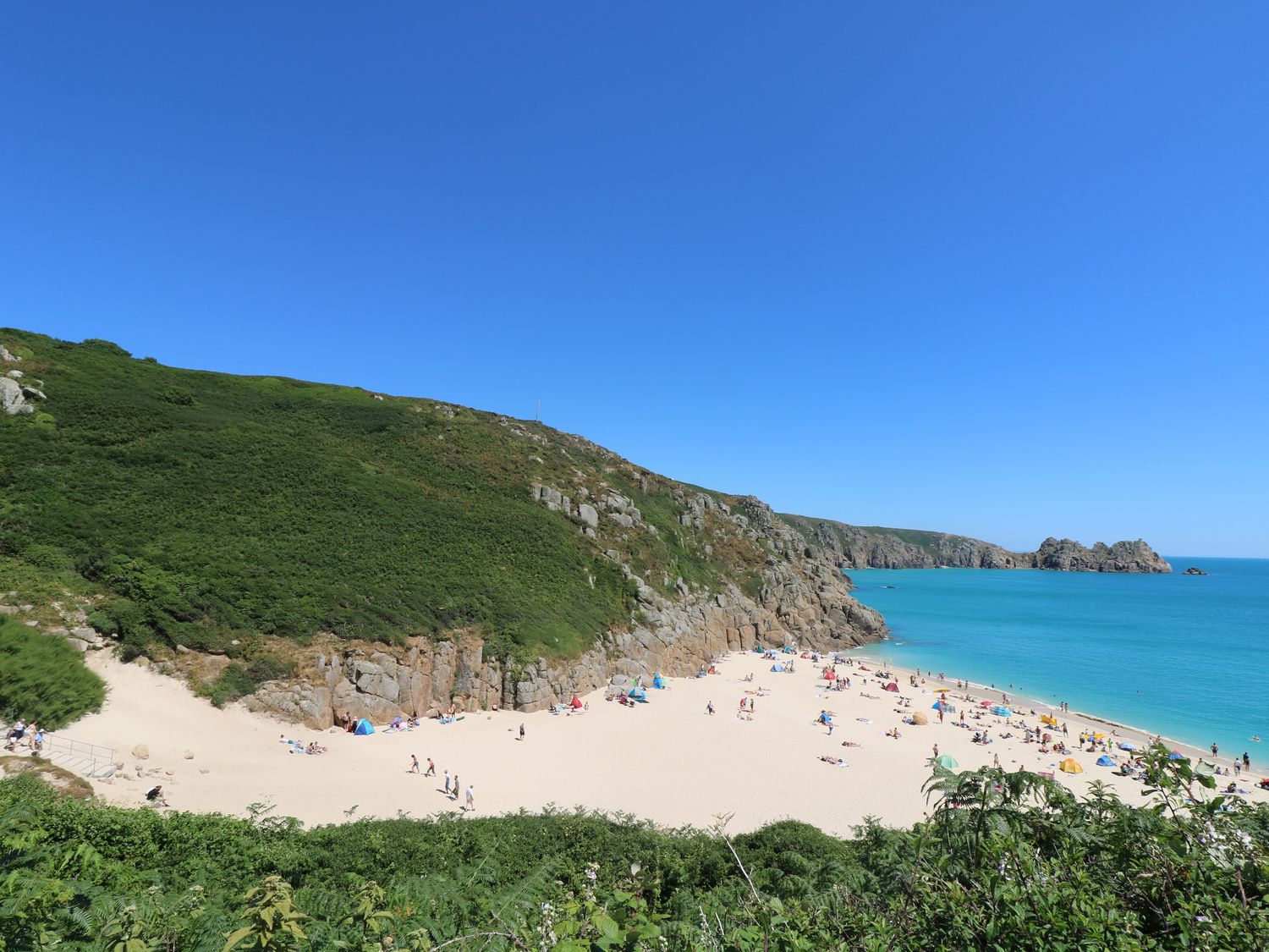A sandy beach with people and umbrellas near rocky cliffs and blue sea at Chynance in Porthcurno
