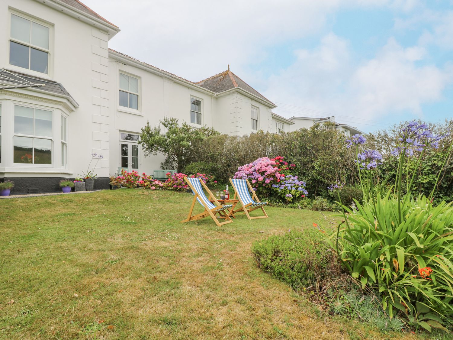 A garden with two wooden deck chairs and a small table in front of a white house with flowers and shrubs at Chynance in Porthcurno