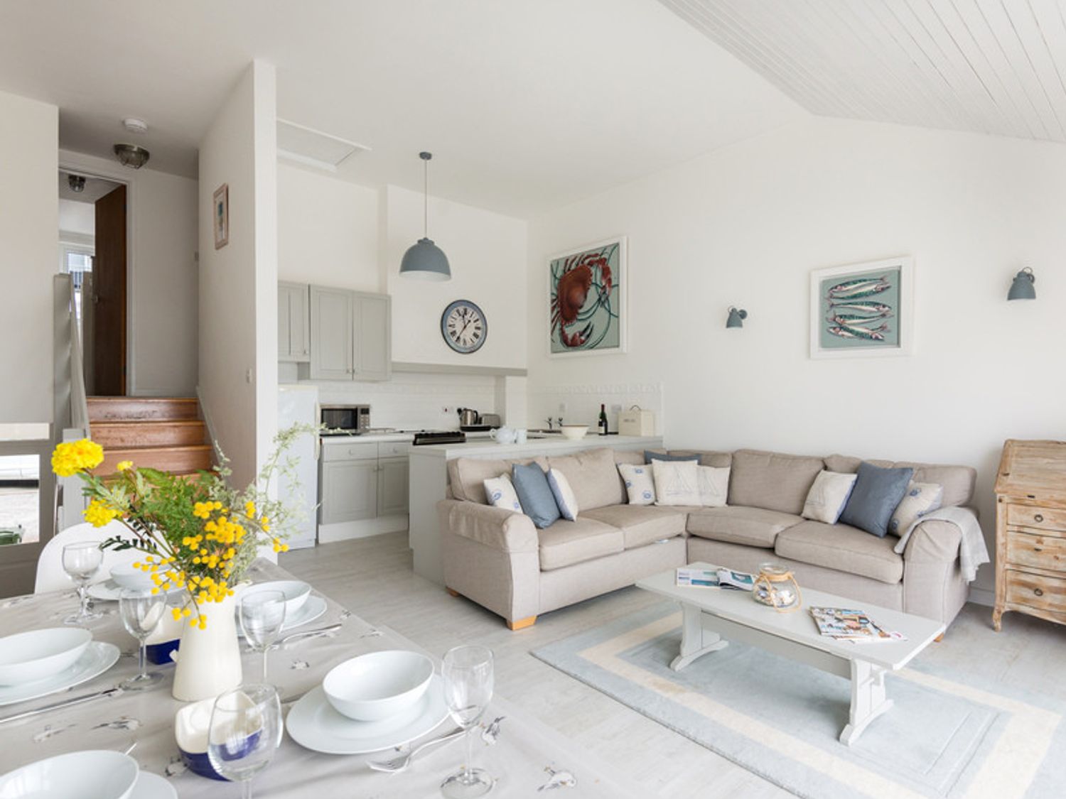 A dining table set with white dishes and a vase of yellow flowers next to a seating area with a beige sectional sofa and a coffee table at Slipway 10 in Rock