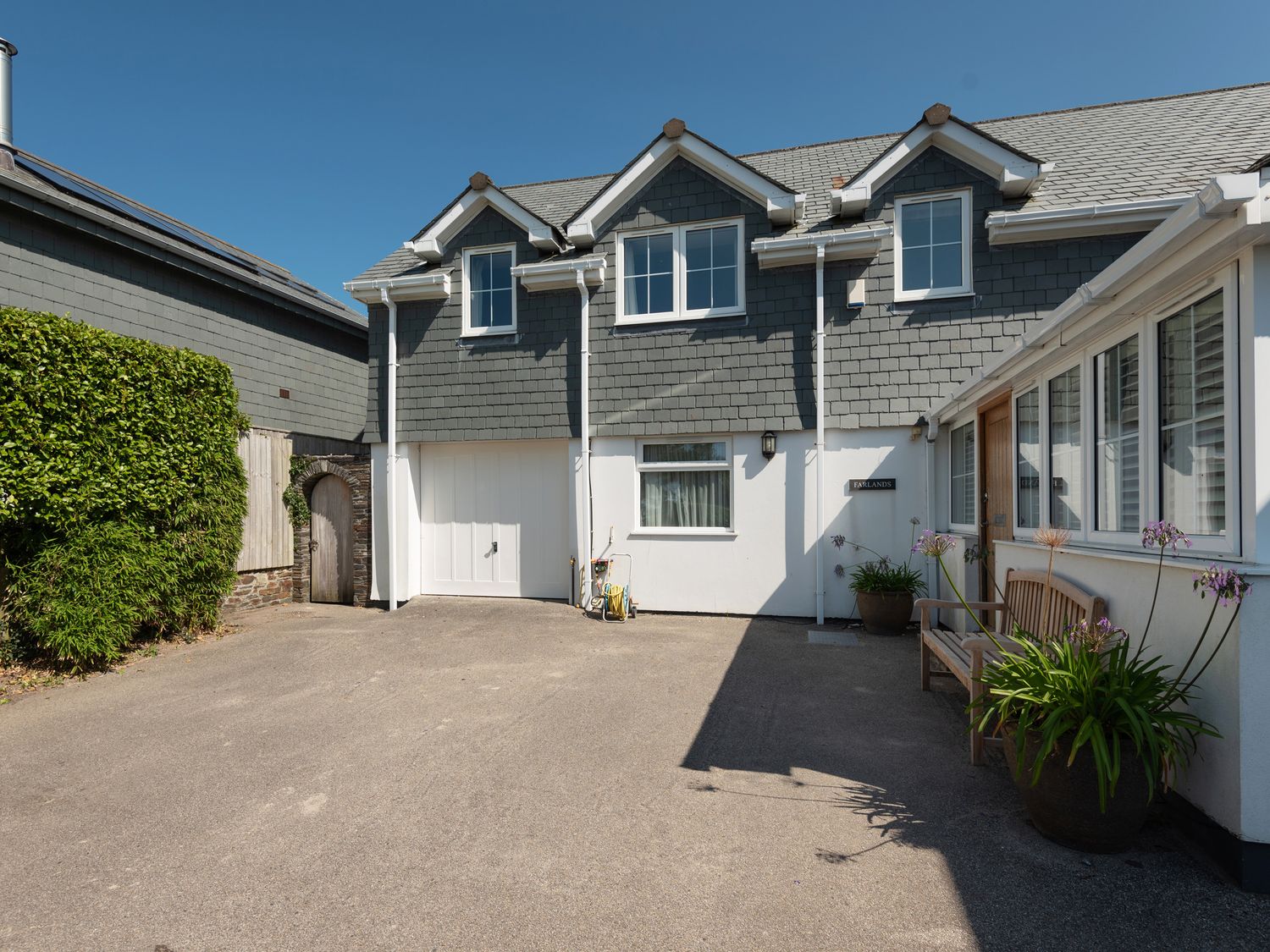 The exterior of a two story house with gray roof tiles white walls and a driveway with plants and a bench at Farlands in Daymer Bay