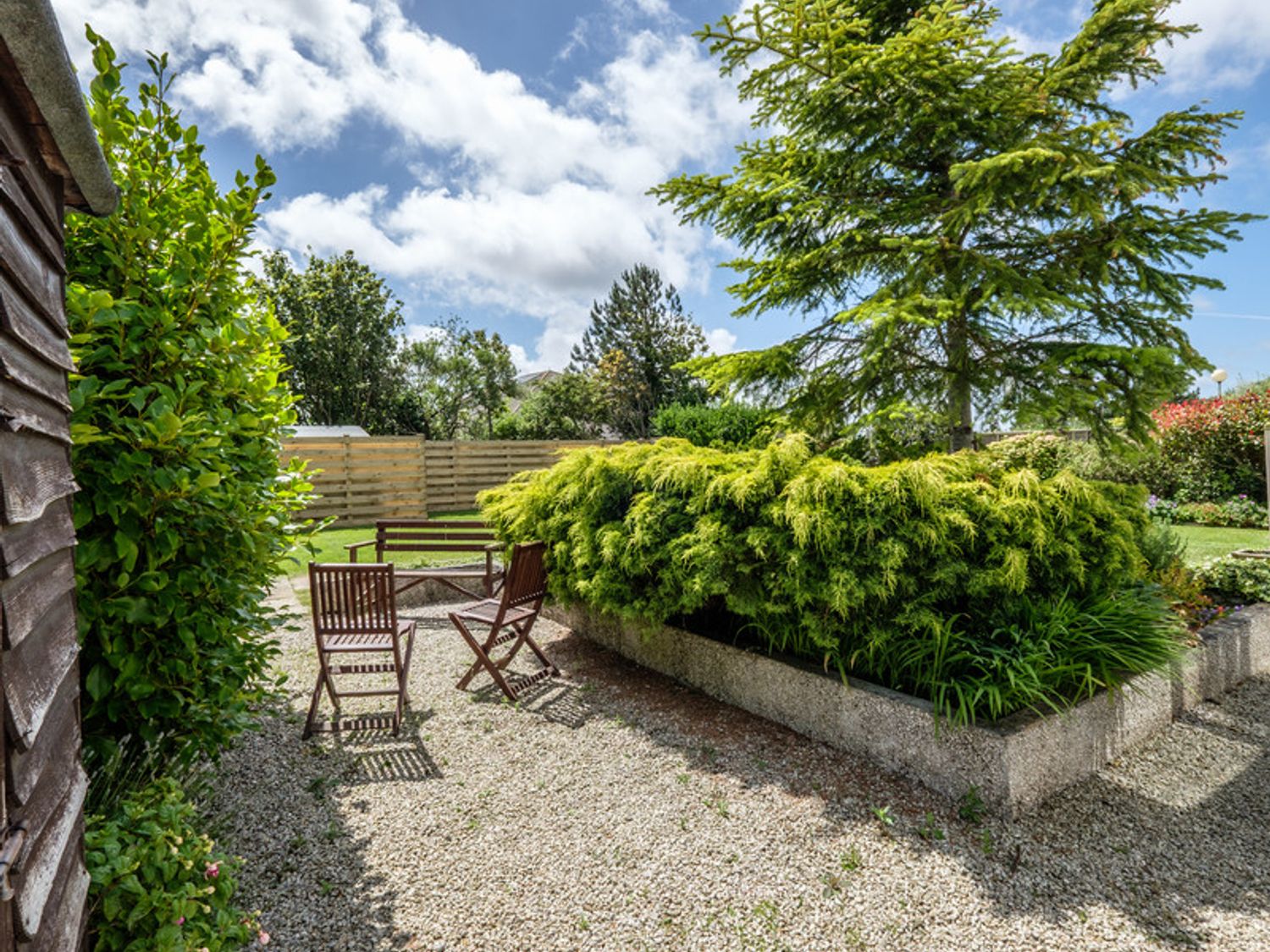 An outdoor garden area with gravel ground wooden chairs a bench and shrubs at Cogenhoe in Rock