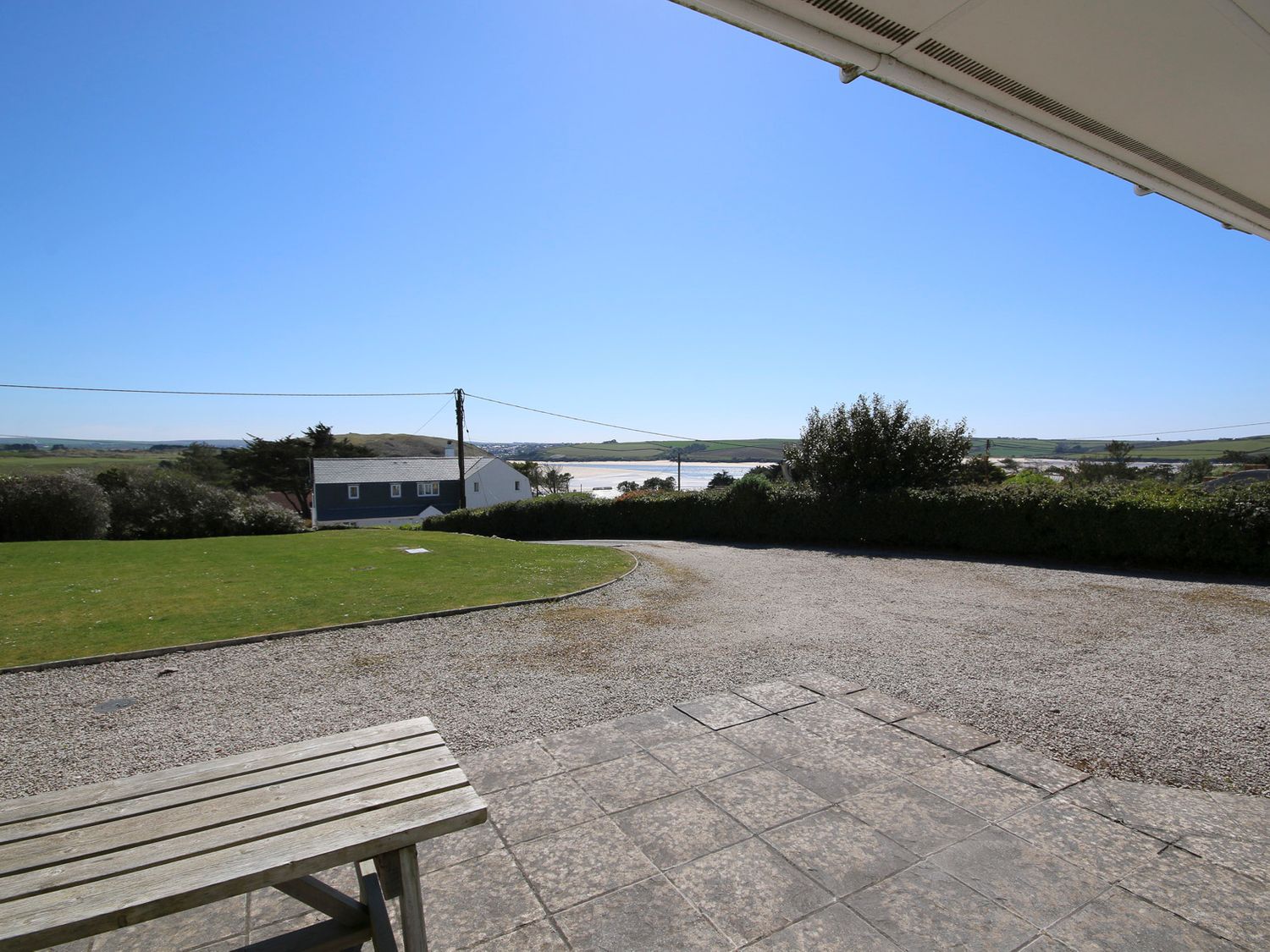 A view of a gravel driveway with a wooden bench and lawn overlooking houses and distant hills at Turnstones in Daymer Bay