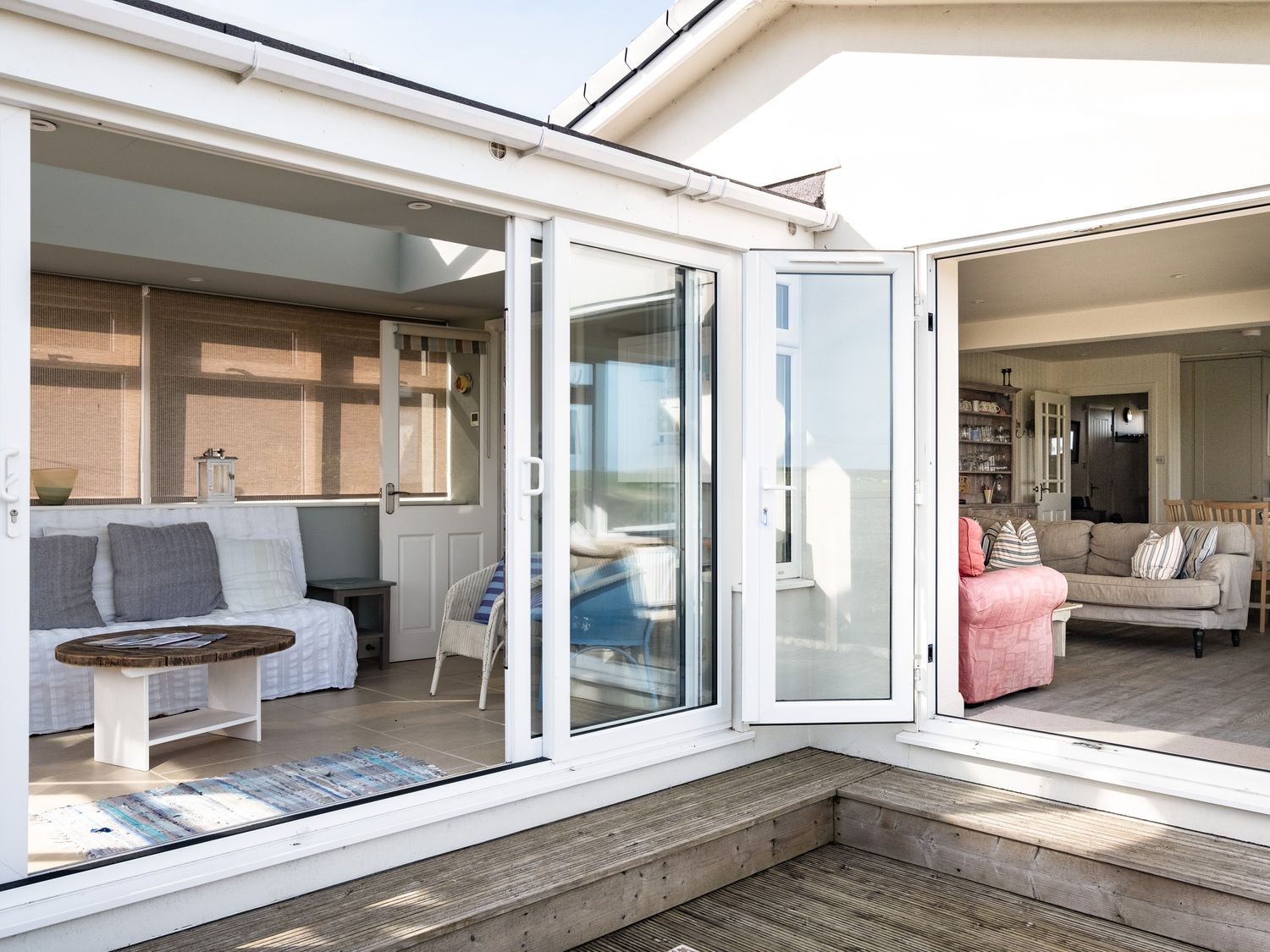 An outdoor wooden deck area connecting two interior rooms with sofas and armchairs at Atlantis in Polzeath